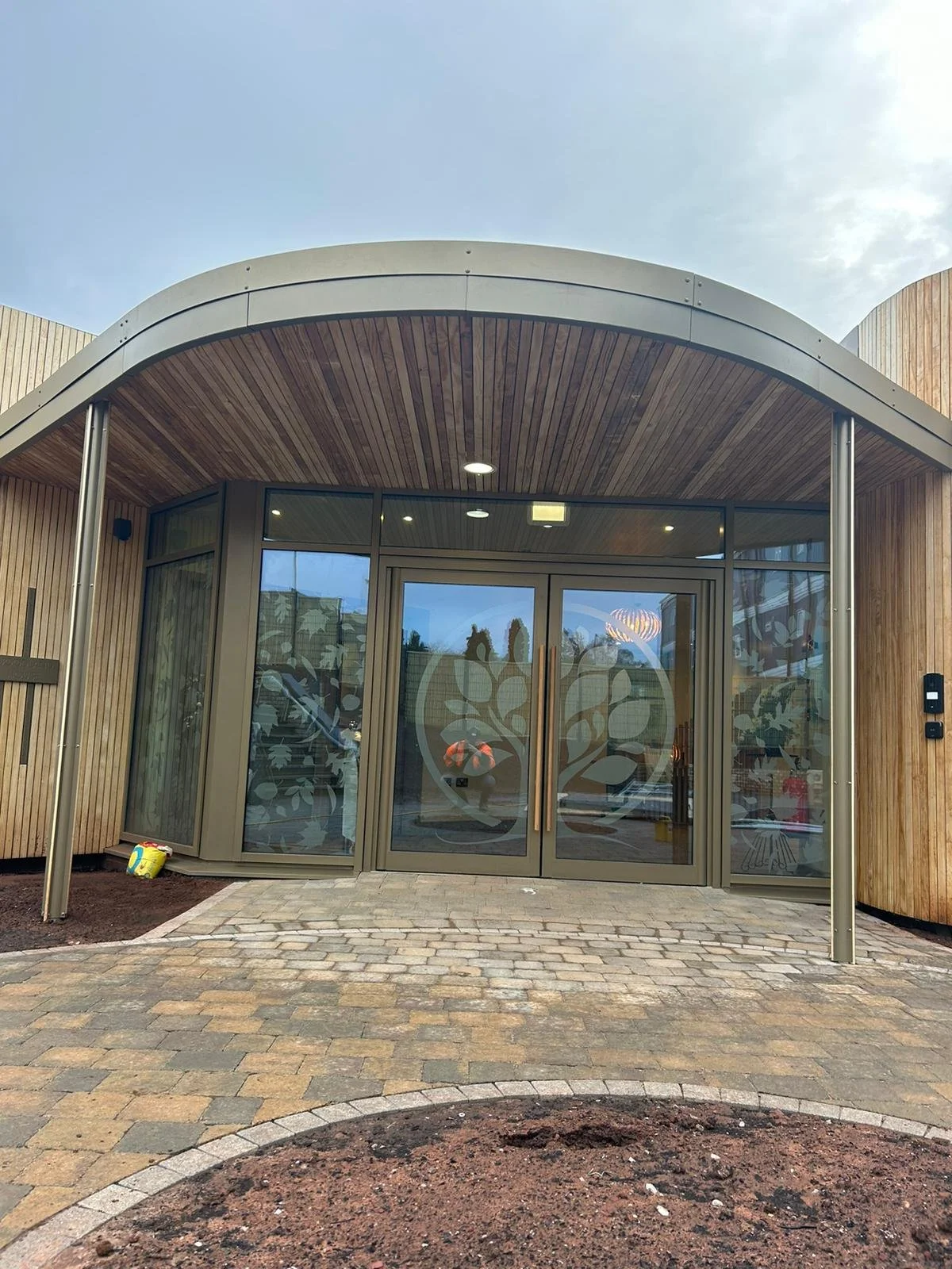 Modern building entrance with glass doors, wooden and metal exterior, and a paved walkway leading up to it.