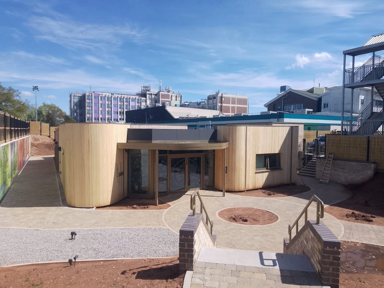 Modern building with curved wooden exterior walls and glass entrance doors, surrounded by newly paved pathways and construction soil, with multi-story buildings in the background under a partly cloudy sky.