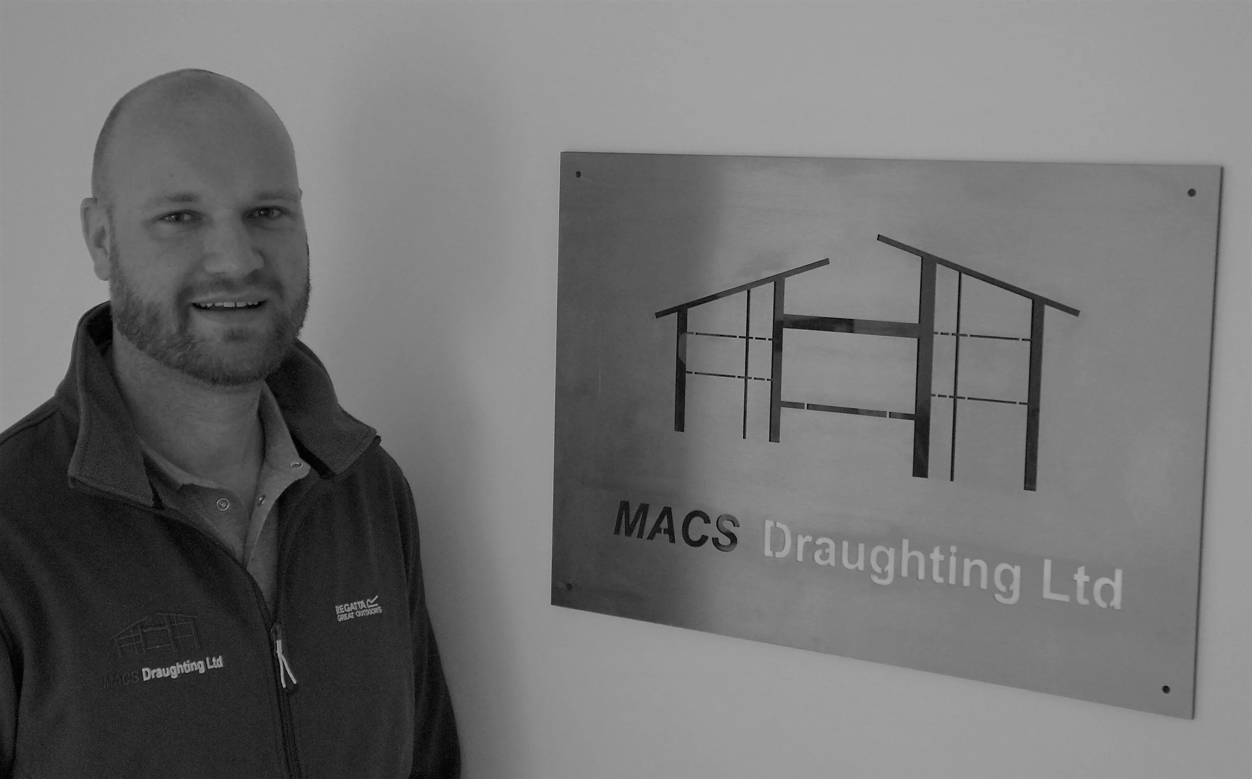A man standing next to a sign that reads "MACS Draughting Ltd" with a simplified building diagram logo.