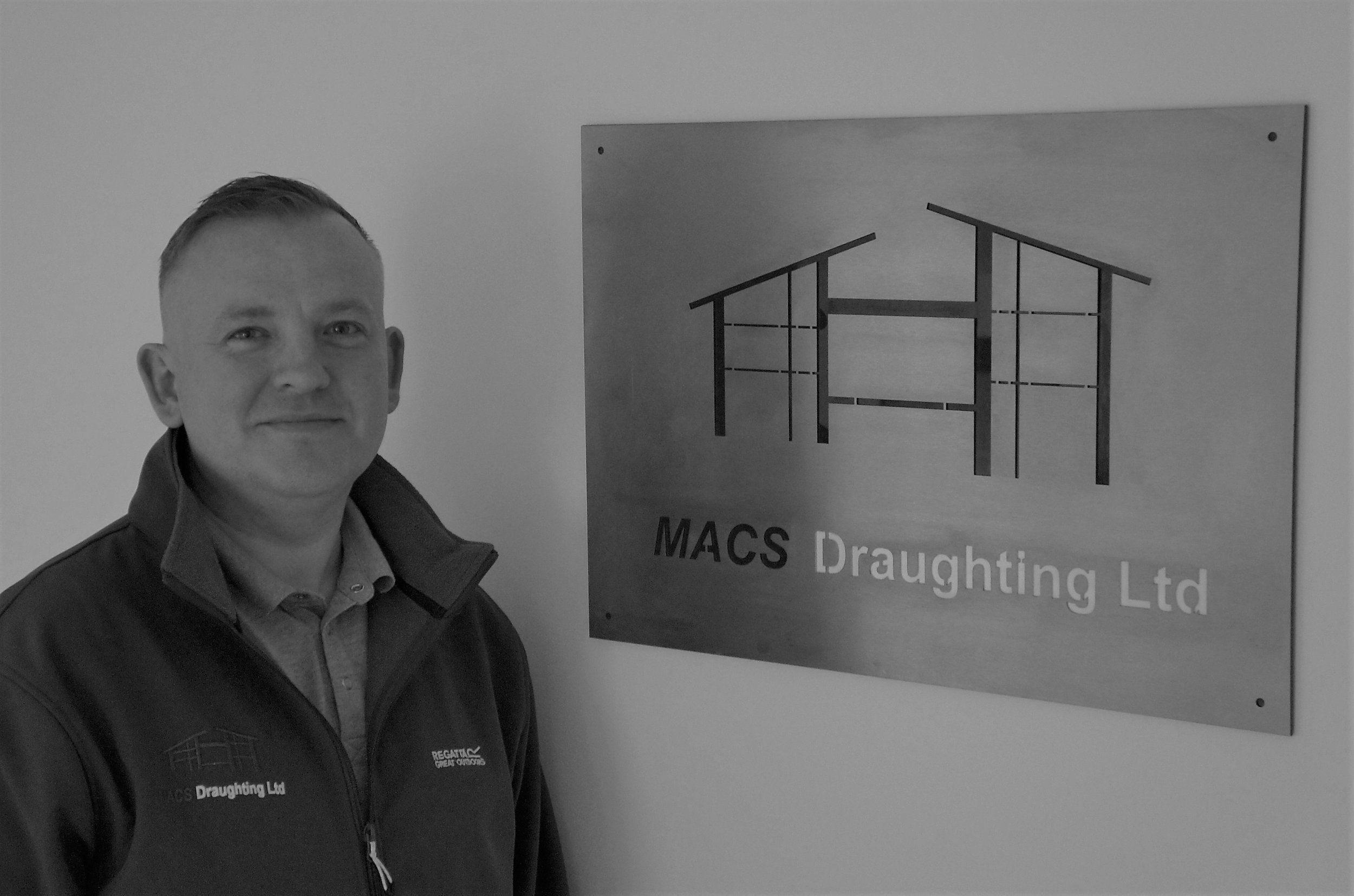 A man in a jacket standing next to a sign for MACS Draughting Ltd, a construction or draughting company.
