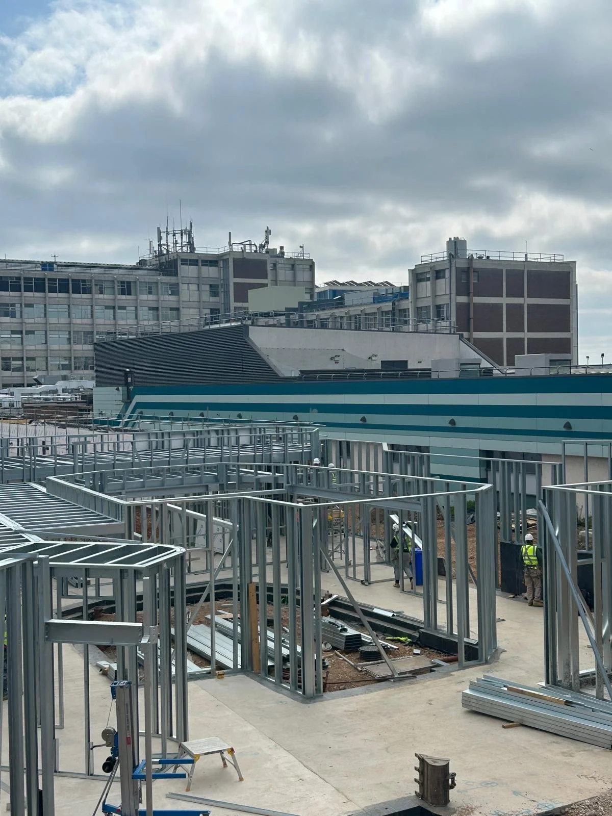 Construction site with metal framework and workers, with a cityscape of office buildings under cloudy sky in the background.