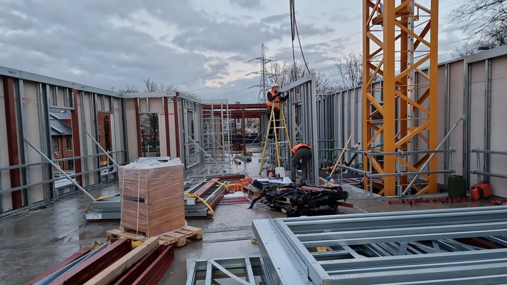 Construction workers building a house with metal framing, with scattered construction materials and a crane in the background.