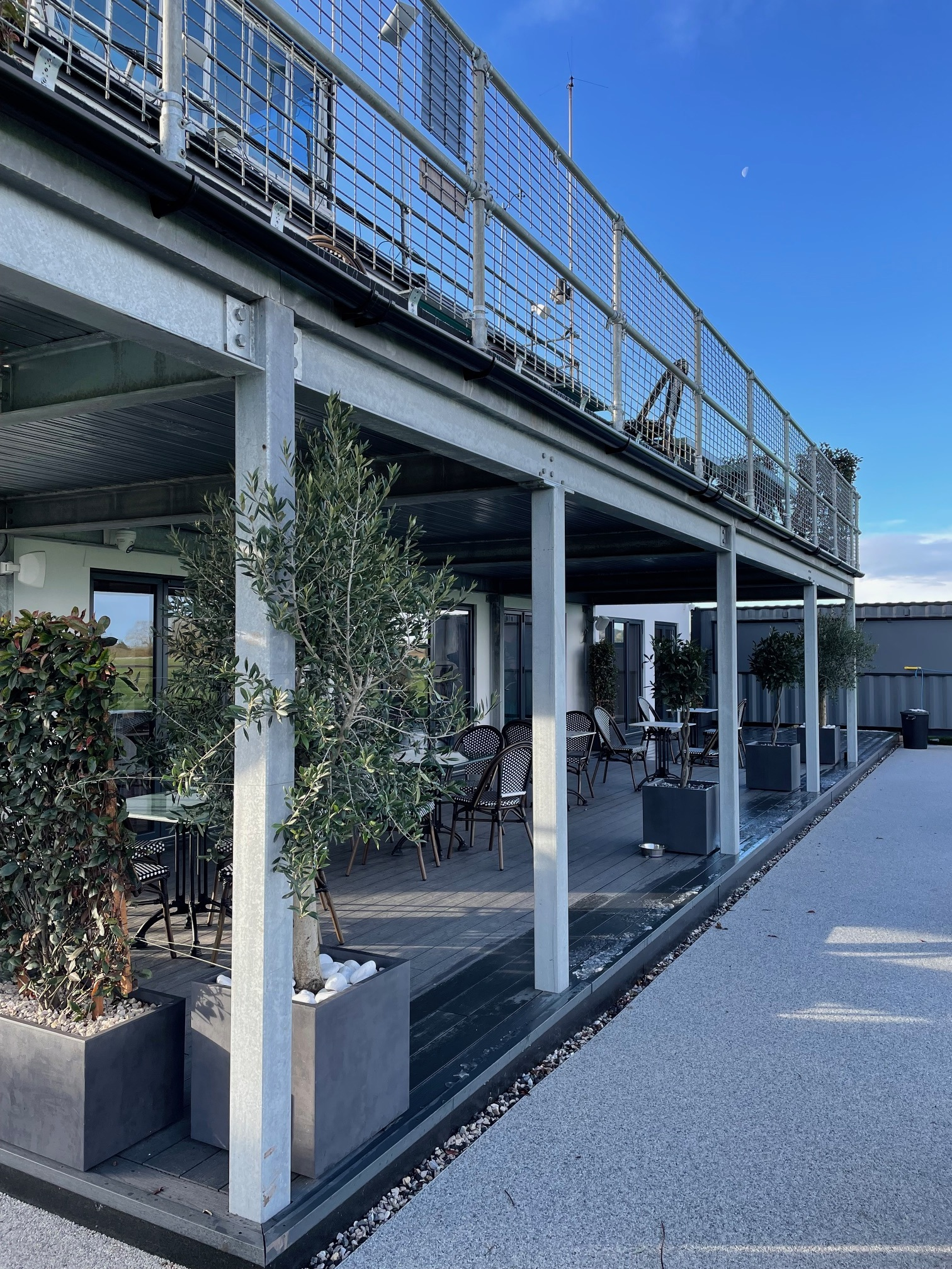 Outdoor seating area with tables, chairs, and potted plants underneath a balcony with metal railing, against a blue sky with visible moon.