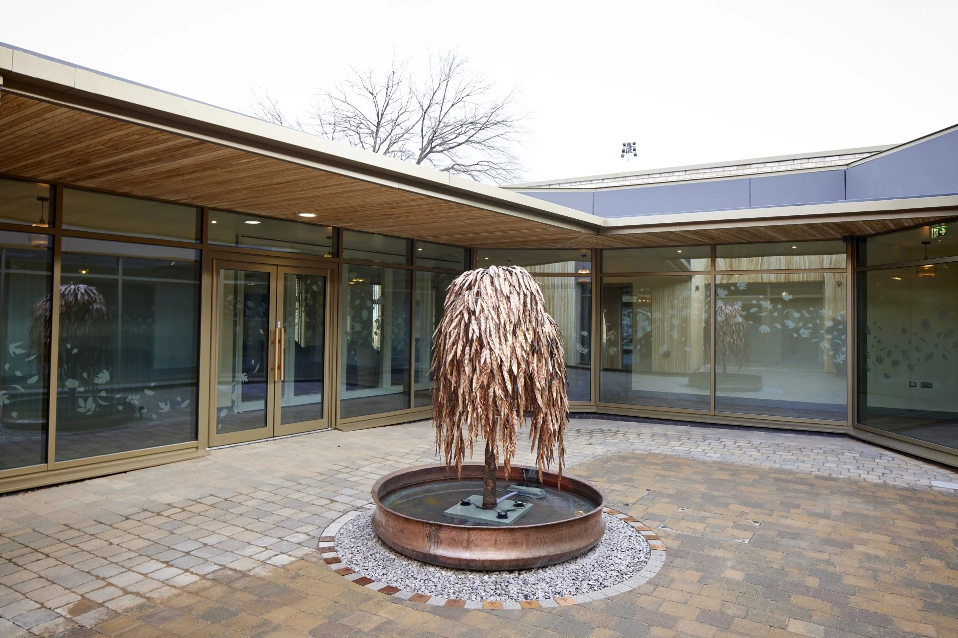 A copper-colored tree-shaped sculpture in a round fountain, set in a paved courtyard surrounded by glass walls of a modern building.