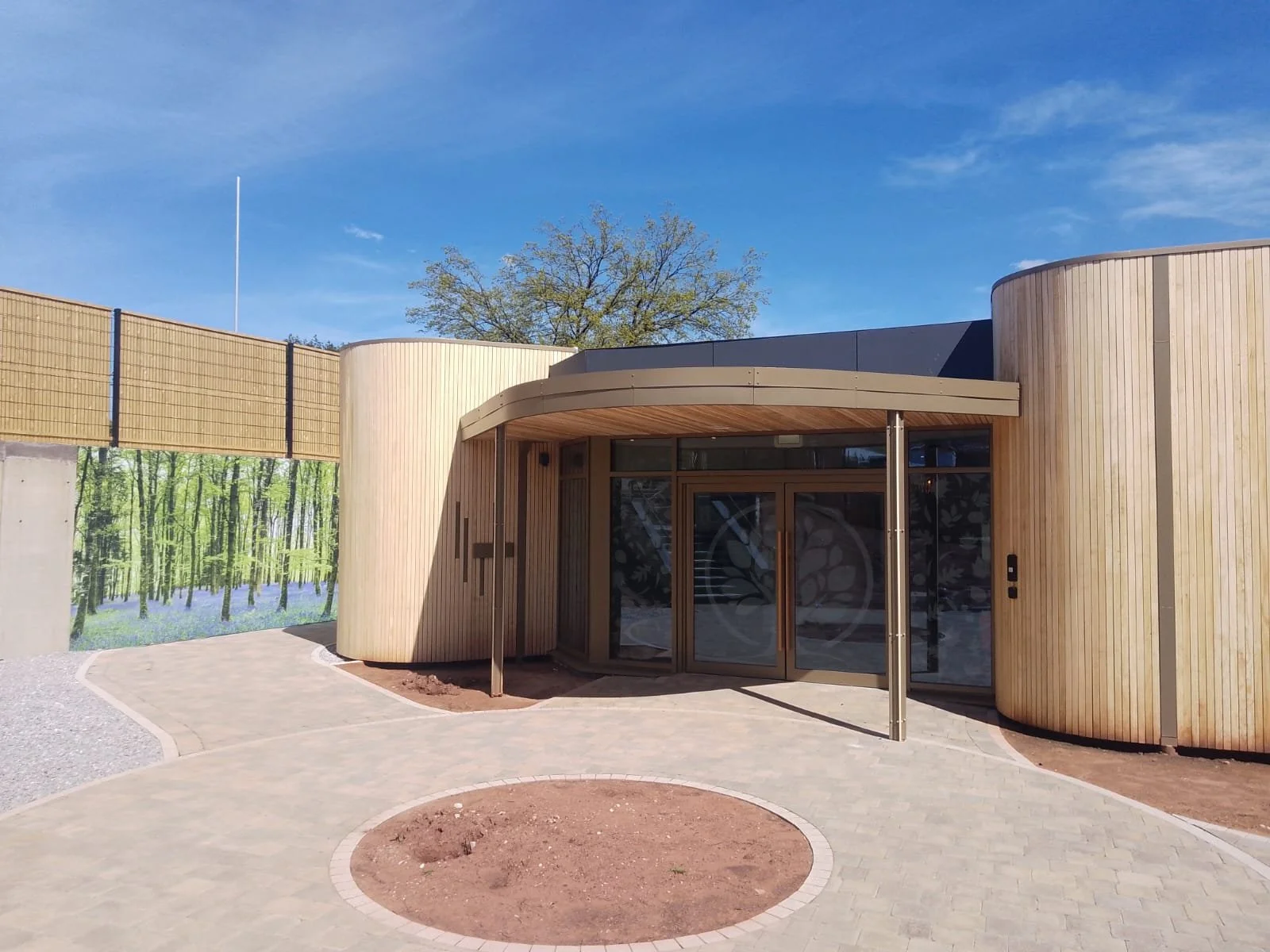 Modern building with curved wooden exterior, glass doors, and a paved walkway leading to the entrance, against a blue sky with some clouds and trees in the background.