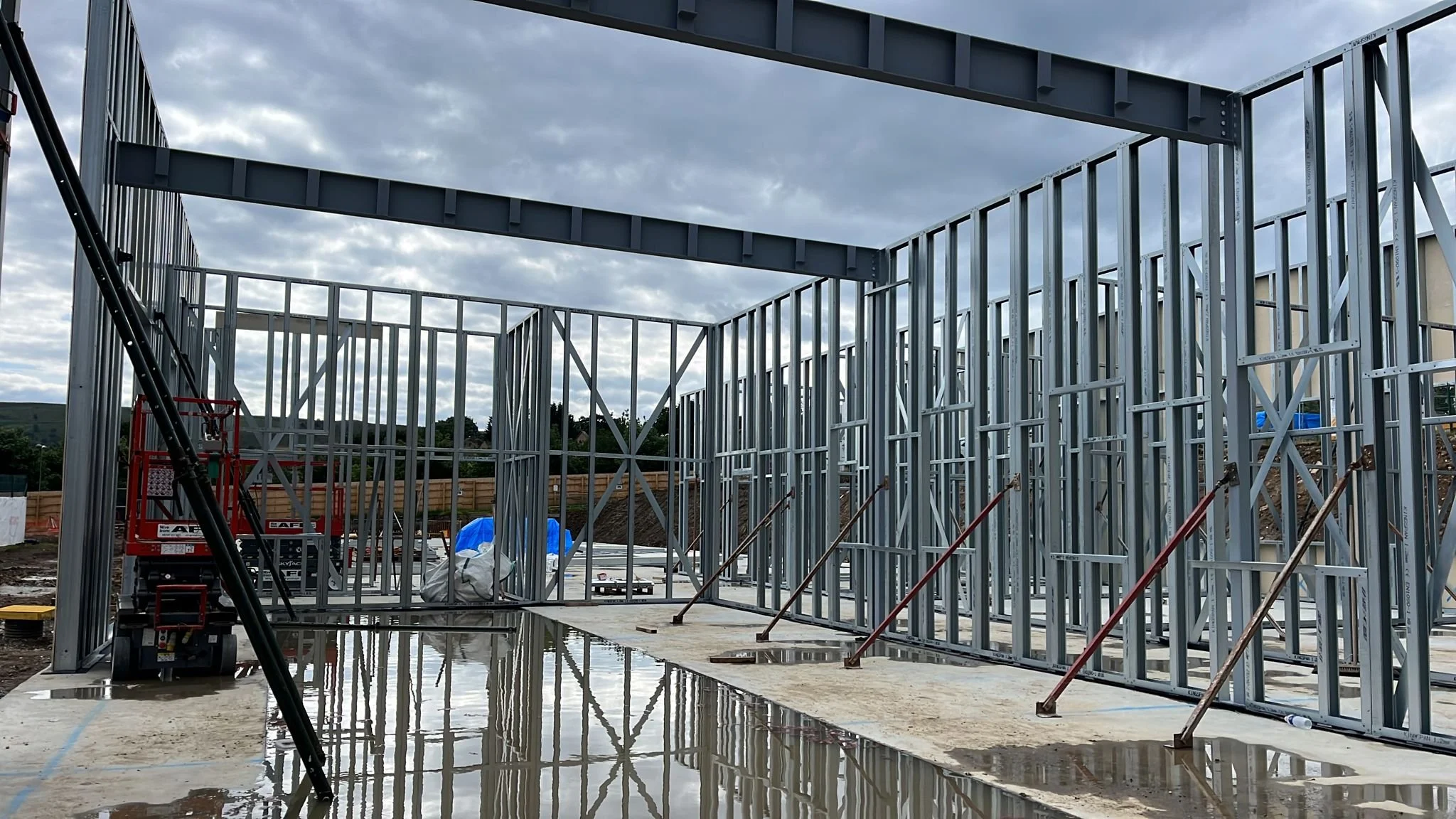 Steel frame structure of a building under construction with a concrete foundation and tools on site, cloudy sky above.