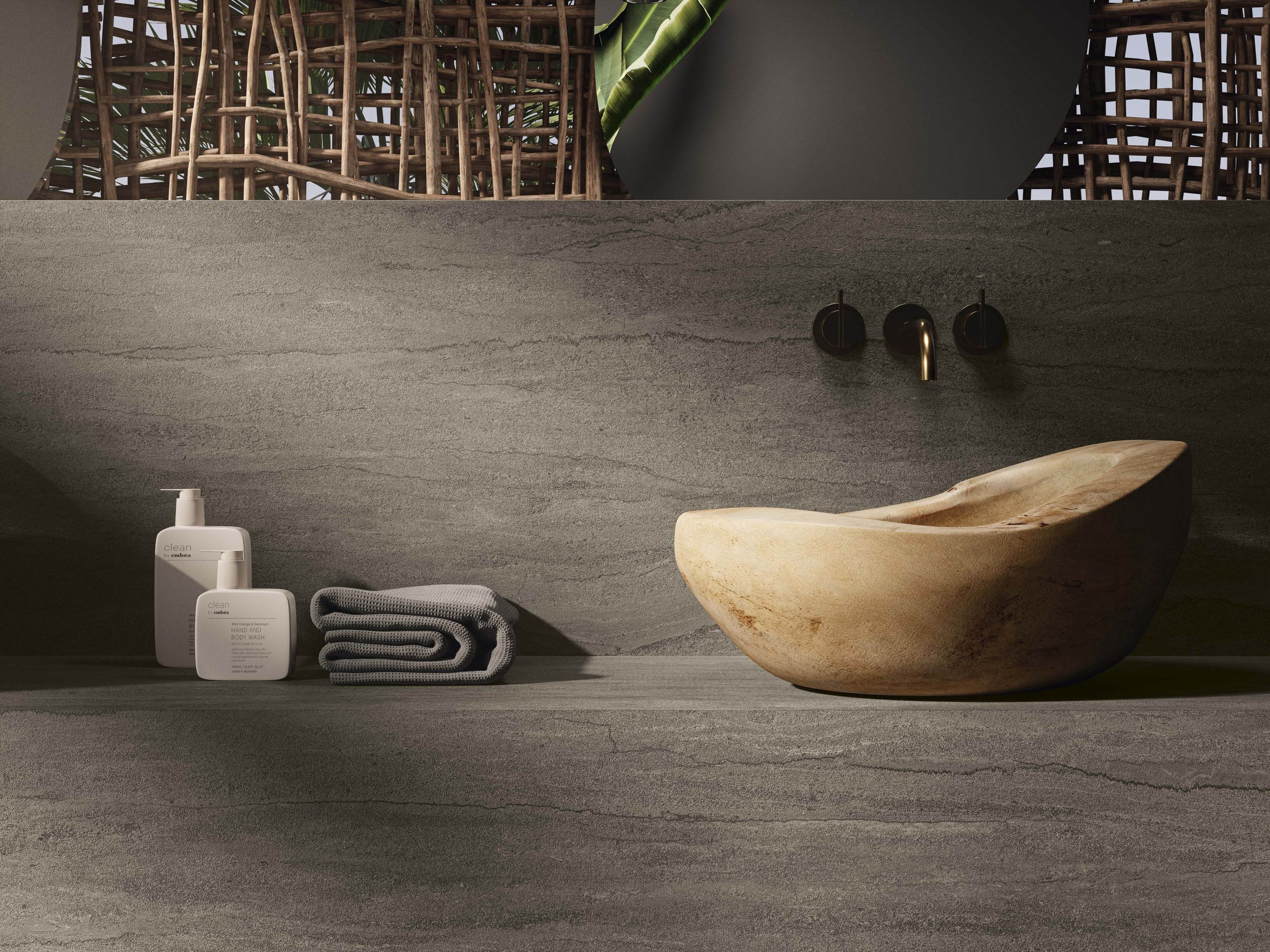 A bathroom countertop with a wooden bowl sink, black wall-mounted faucet, and amenities including soap and hand wash, along with a folded gray towel. In the background, there are decorative woven wooden partitions and green leaves.