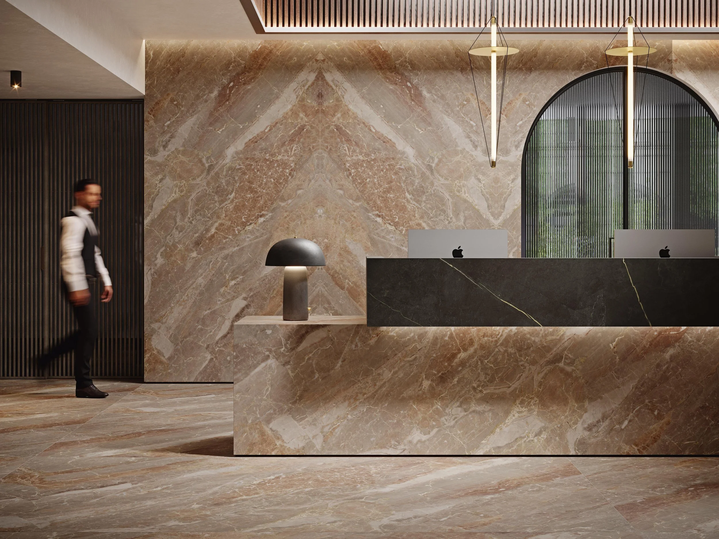 Modern hotel reception area with marble walls and floor, a black marble countertop with Apple computers, a decorative black table lamp, and gold pendant lights, with a blurred man walking past.