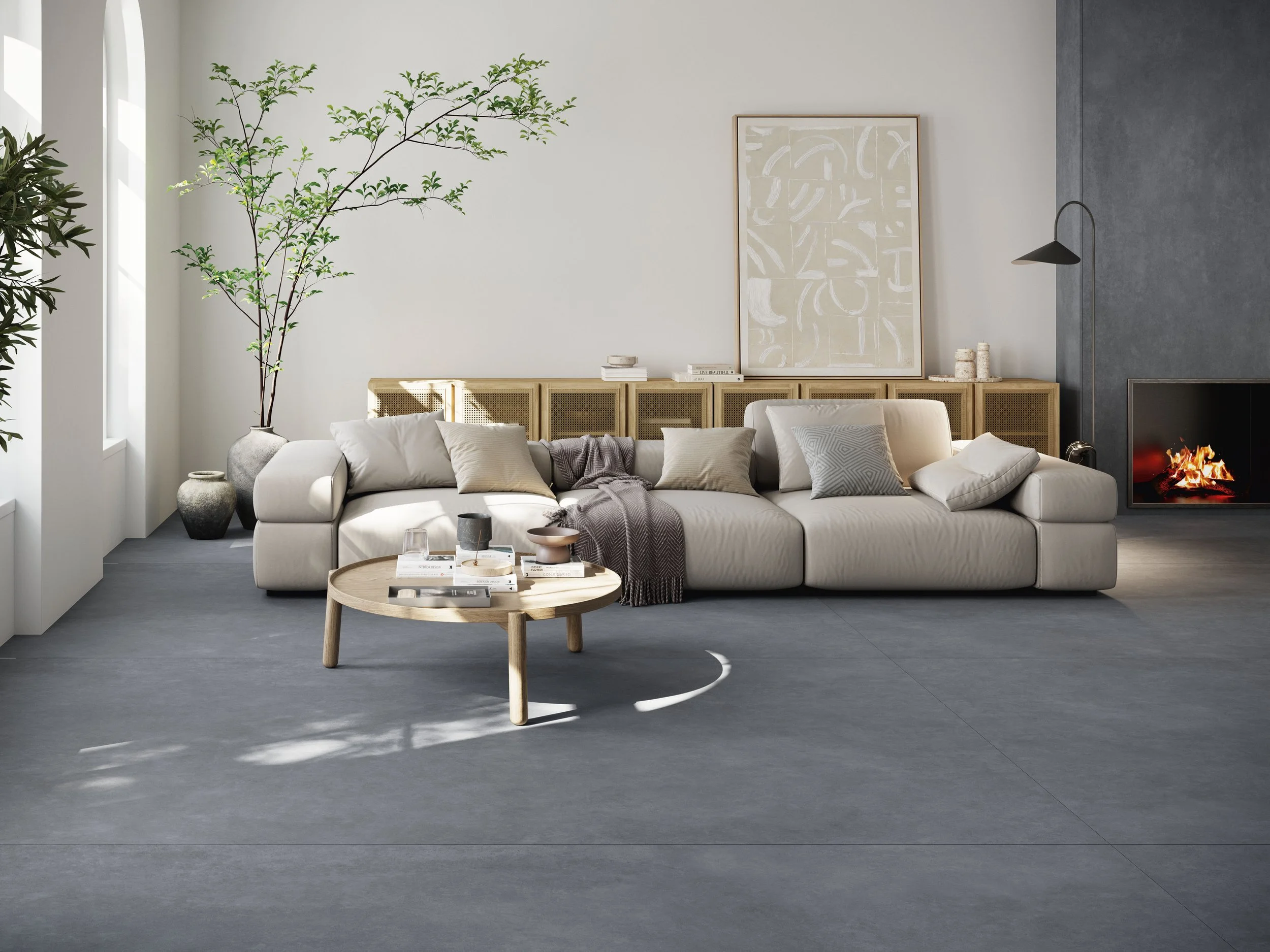 Modern living room with a white sectional sofa, multiple throw pillows, a round wooden coffee table with books and decor, large potted plants, a fireplace with a fire, and minimalist artwork on the wall.