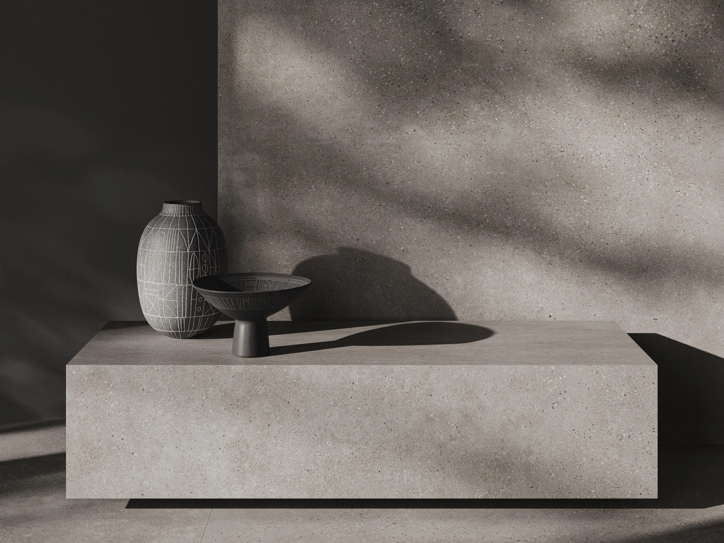 A decorative arrangement featuring a tall, gray vase with geometric line patterns, a shallow black bowl with similar line patterns on a pedestal, all on a rectangular, beige stone platform with textured wall behind.