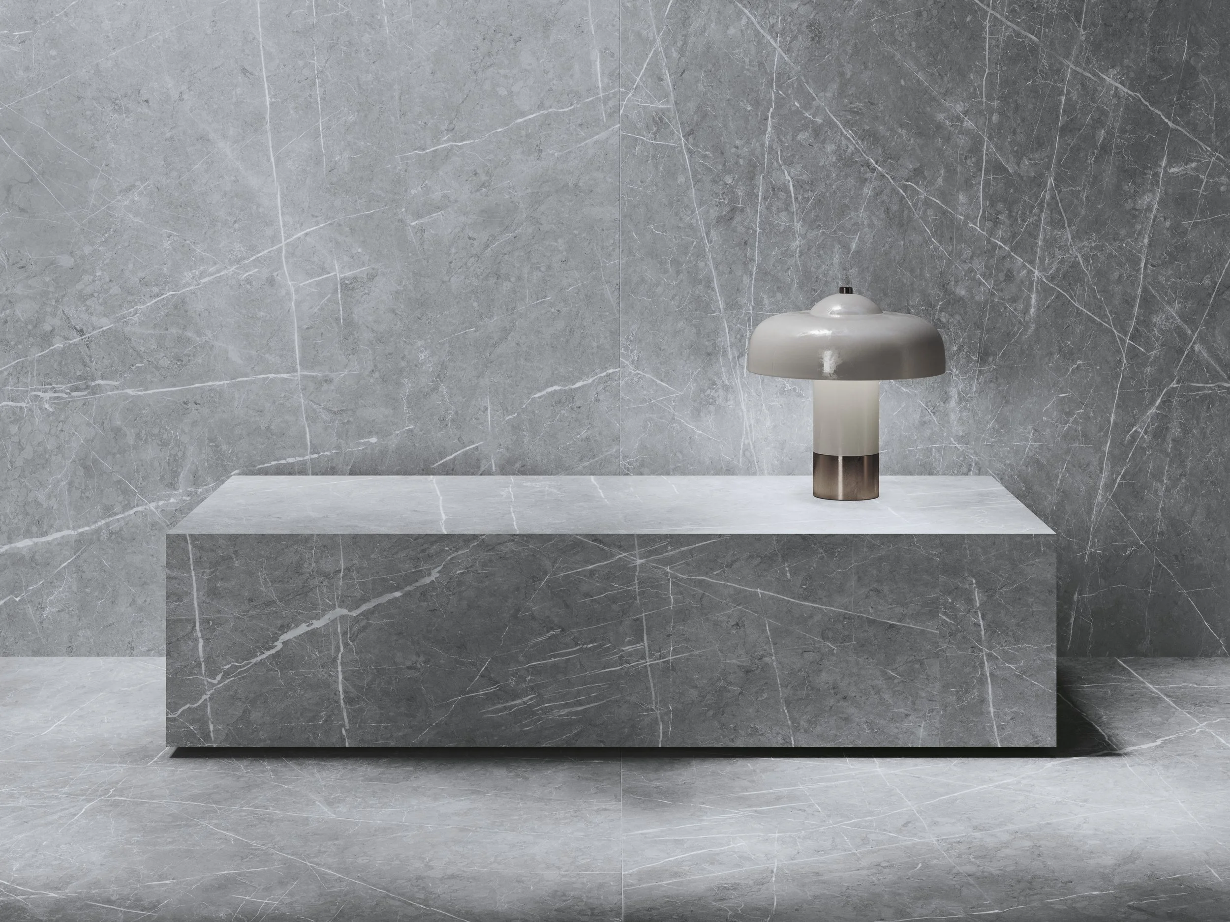 A sleek modern table lamp with a rounded shade sitting on a minimalist marble platform against a grey marble wall and floor.