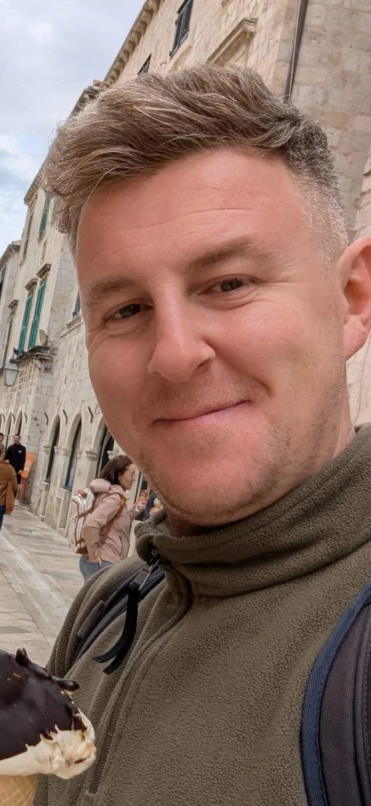 A man with short, light brown hair and slight stubble, wearing a pink t-shirt, looking at the camera against a bright blue background.