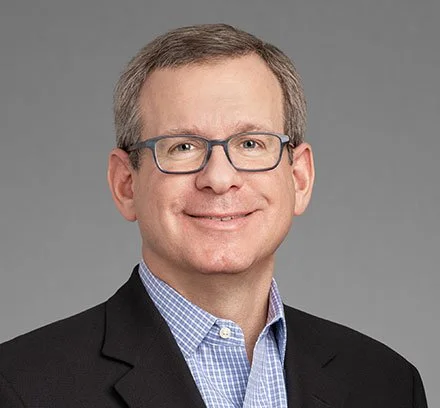 Professional headshot of a smiling man with glasses, wearing a black suit jacket and blue checkered shirt, against a gray background.