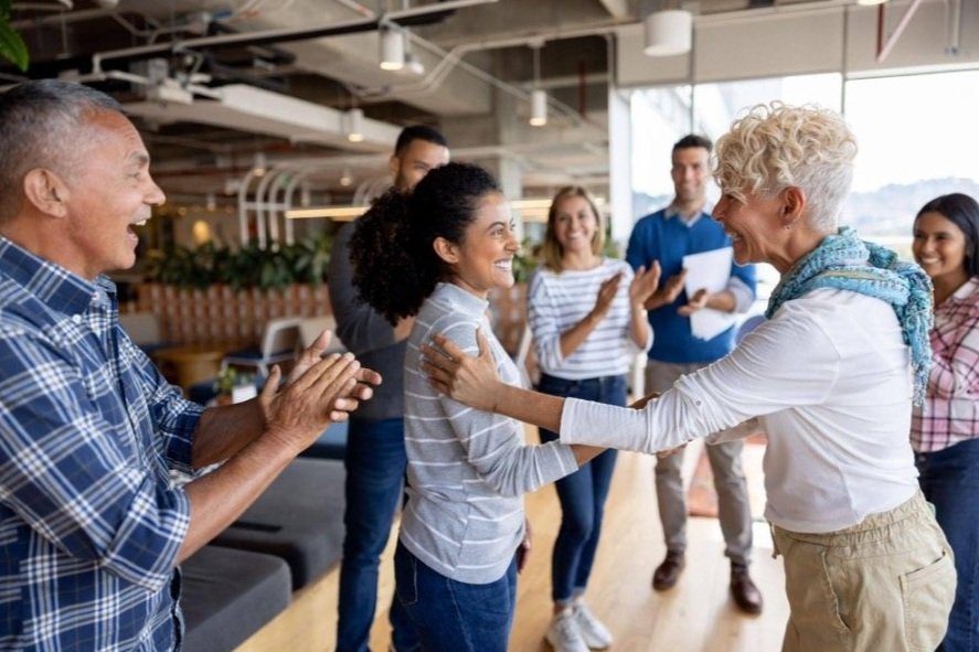 Group of diverse people in a modern, well-lit office celebrating, some clapping and smiling, with two women in the center exchanging a warm hug.