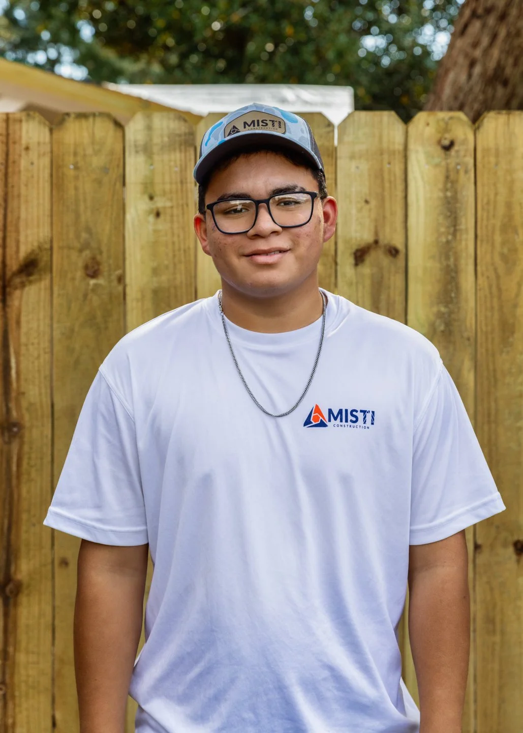 A young man wearing glasses, a baseball cap, and a white t-shirt with a construction company logo, standing outdoors in front of a wooden fence.