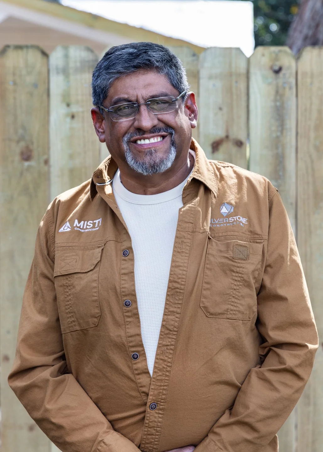 Smiling man wearing a dark suit, white shirt, orange tie, blue reflective sunglasses, and a lapel pin, with a bright blue sky in the background.