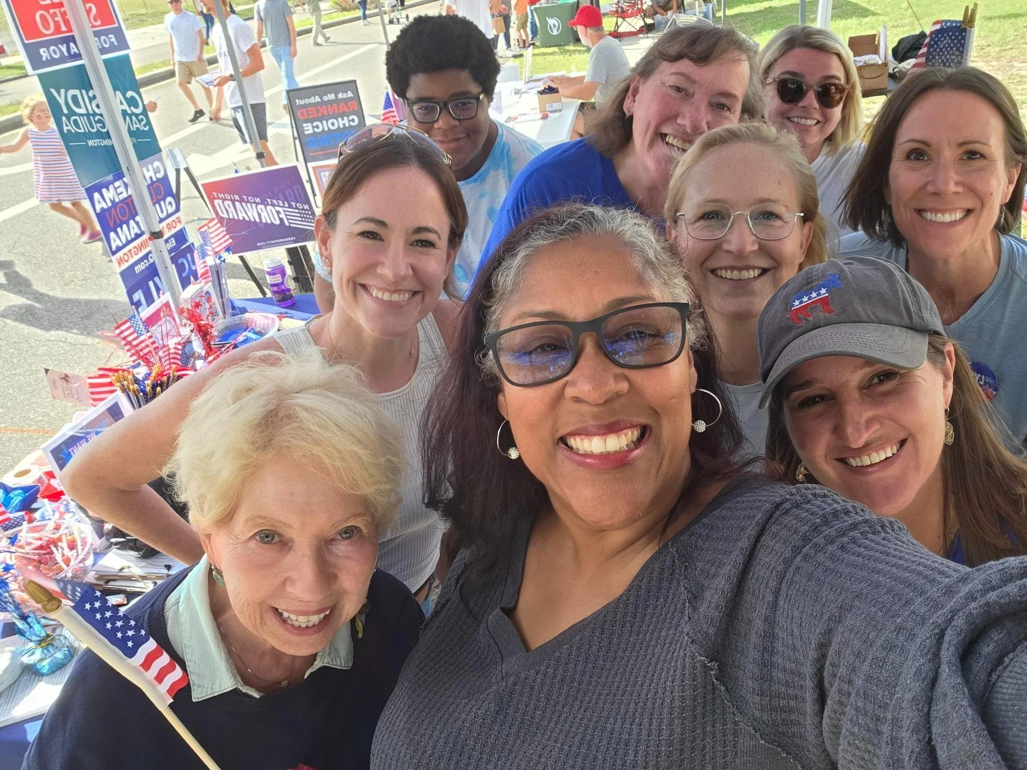 A group of people smiling for a selfie at a political event with campaign signs and patriotic decorations in the background.