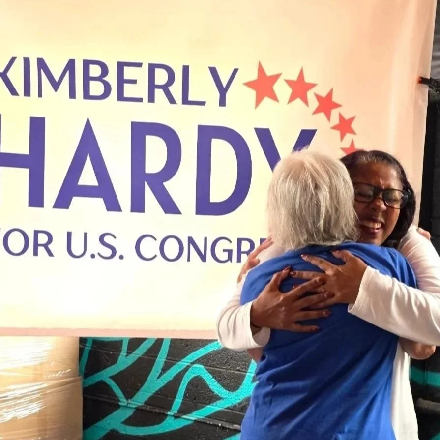 Two women hugging at a political campaign event with a sign that reads 'Kimberly Hardy for U.S. Congress' in the background.