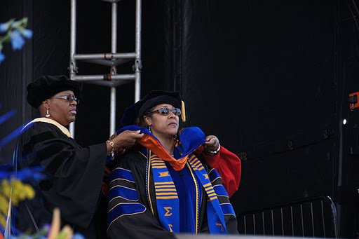 Two women in graduation robes and caps, one adjusting the other's hood, on stage at a graduation ceremony.