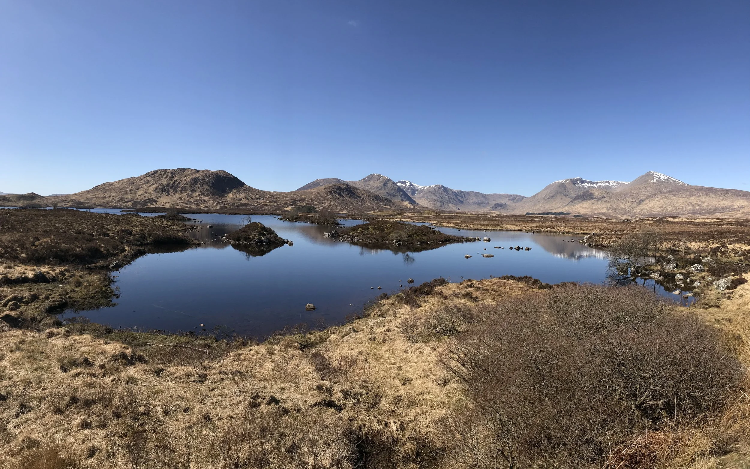Panoramic photograph of a heart-shaped loch reflecting the deep blue sky, surrounded by snow-capped mountains, with a smaller inverted heart shape visible in the foreground grasses.