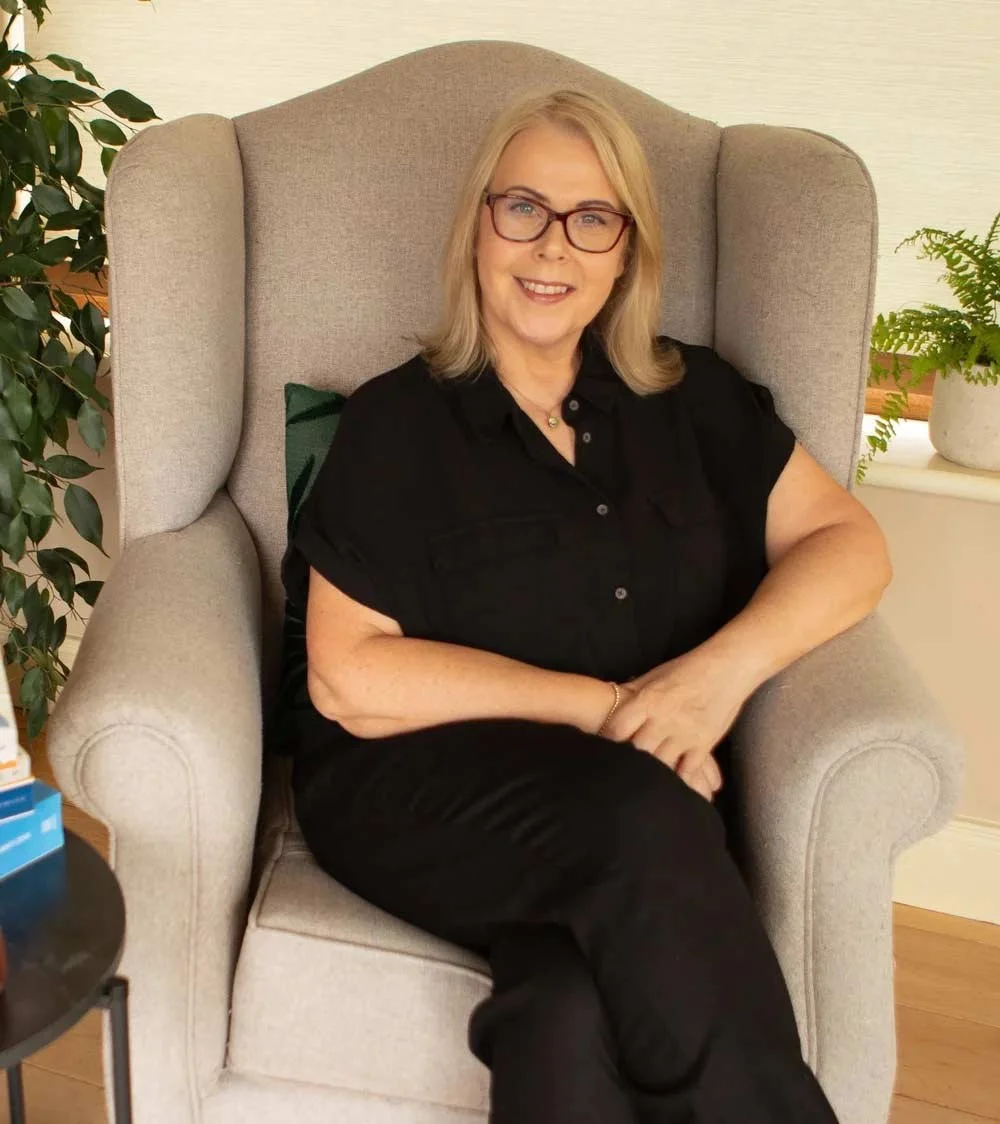 A woman with blonde hair and glasses sitting on a beige armchair, smiling at the camera, in a room with plants and a side table.