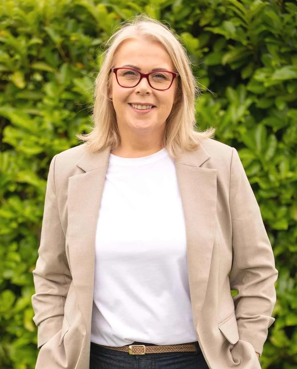 A woman with blonde hair, wearing glasses, a light beige blazer, and a white shirt, stands outdoors in front of green shrubbery, smiling at the camera.