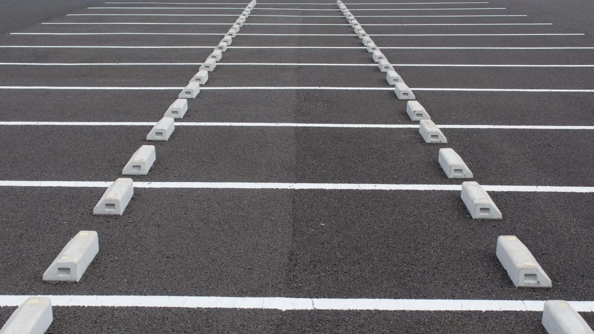 An empty parking lot with a few people walking, surrounded by stores and buildings under an overcast sky