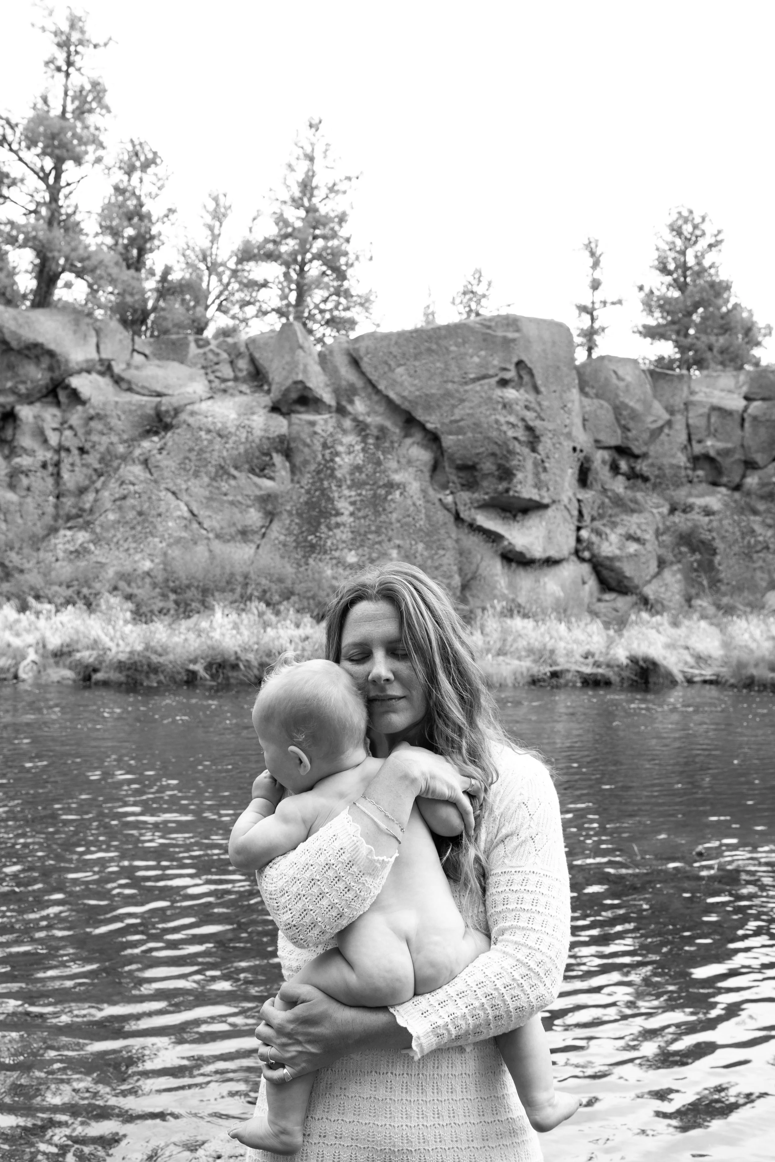 A woman with long hair holding a young child, both of them are naked, standing in a body of water with rocks and trees in the background.