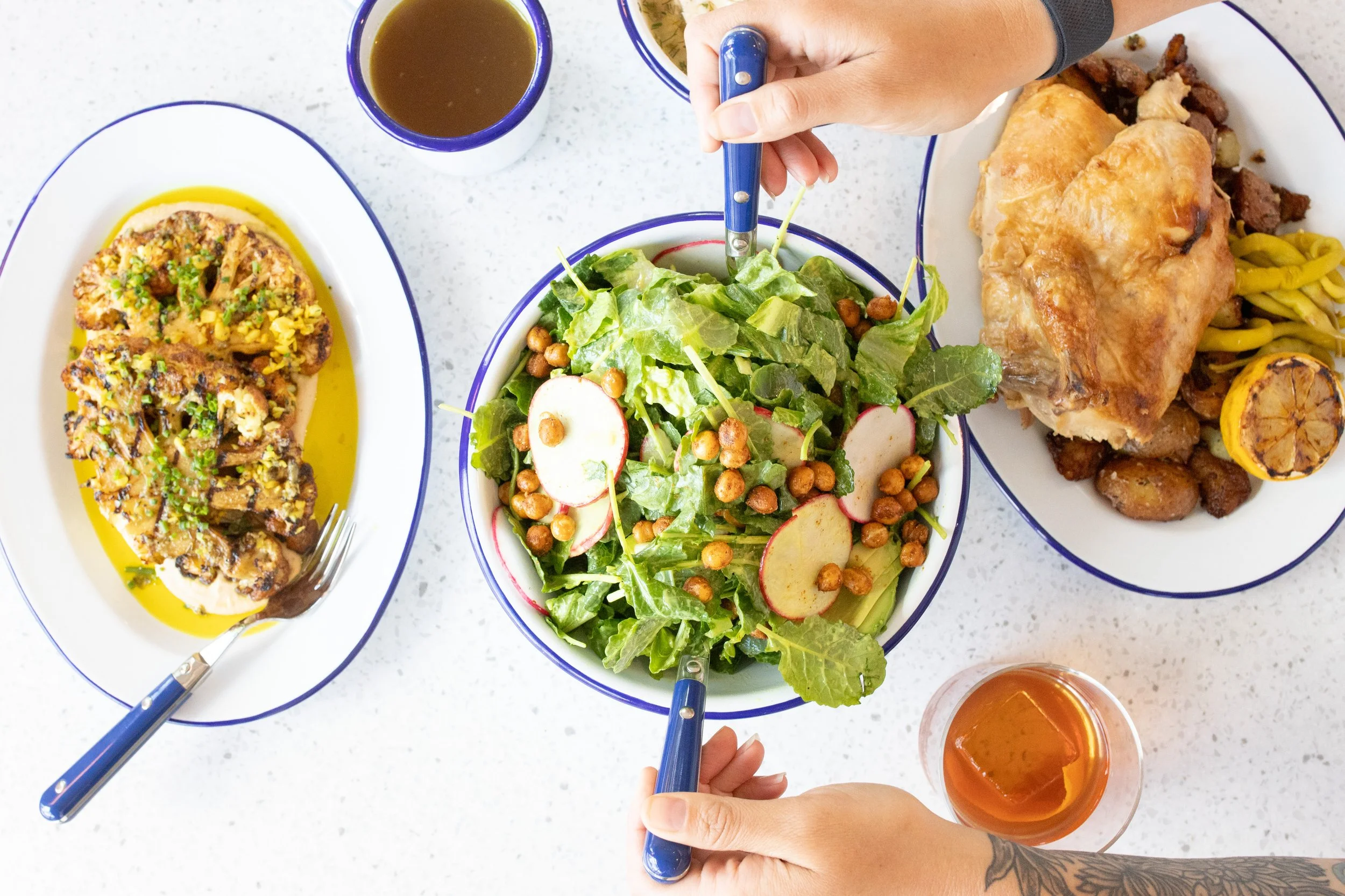 Overhead view of a meal including a colorful salad with radishes and chickpeas, a roasted chicken with lemon, a breaded dish, and various drinks, with two hands holding forks over the salad.