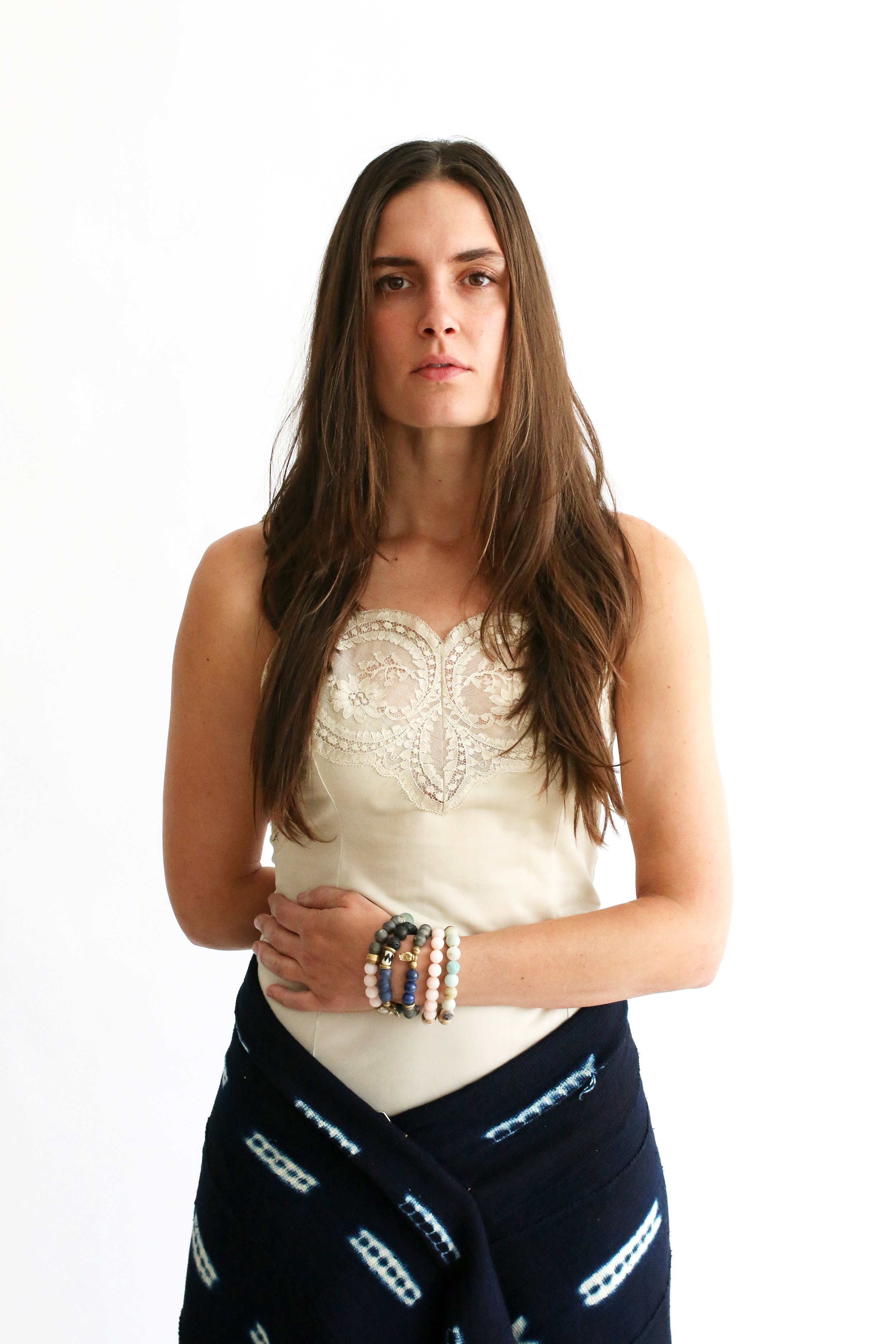 A woman with long brown hair standing against a plain white background, wearing a cream-colored sleeveless top with lace detail, and a dark patterned skirt. She has multiple beaded bracelets on her left wrist and one on her right wrist, and she looks