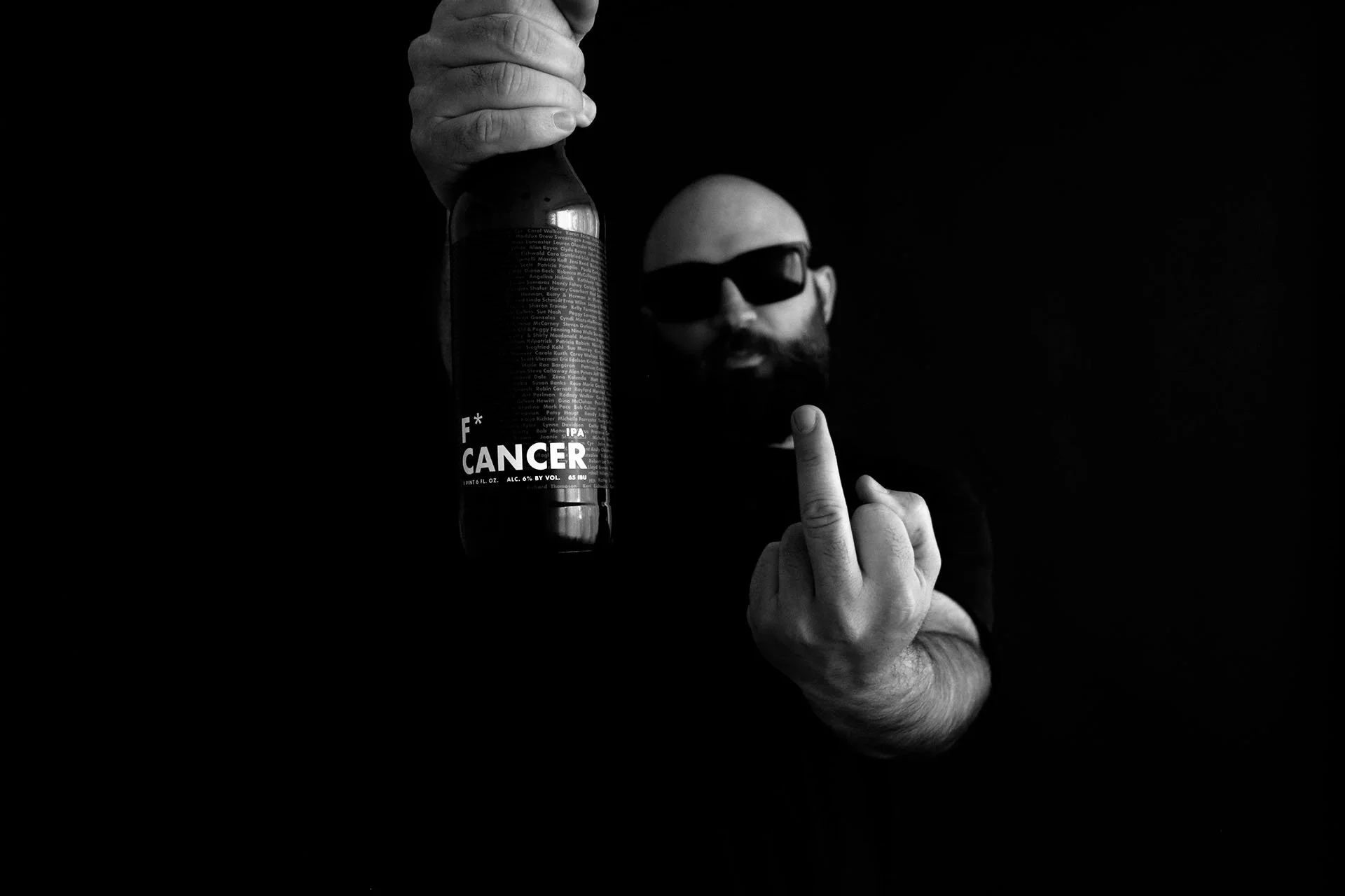 A man with a bald head and beard wearing sunglasses holds a dark bottle labeled 'F* Cancer' and shows his middle finger.