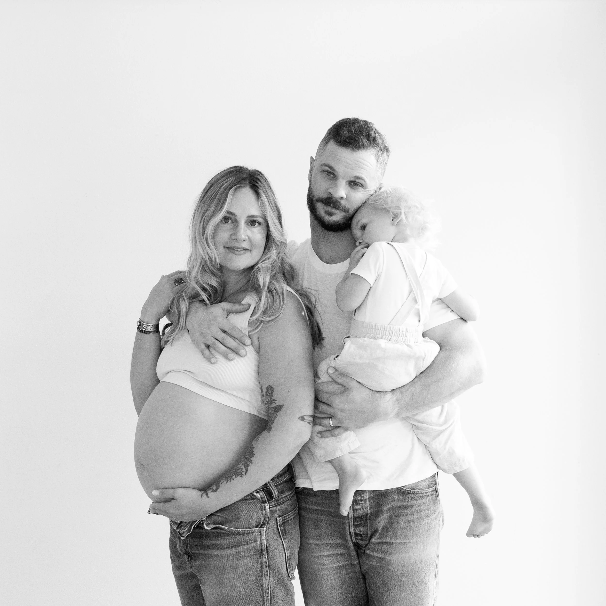 Black and white photo of a pregnant woman, a man, and a young child in front of a plain background. The woman is smiling, holding her pregnant belly, and has a tattoo on her arm. The man is holding the child, who is resting on his shoulder, and the c