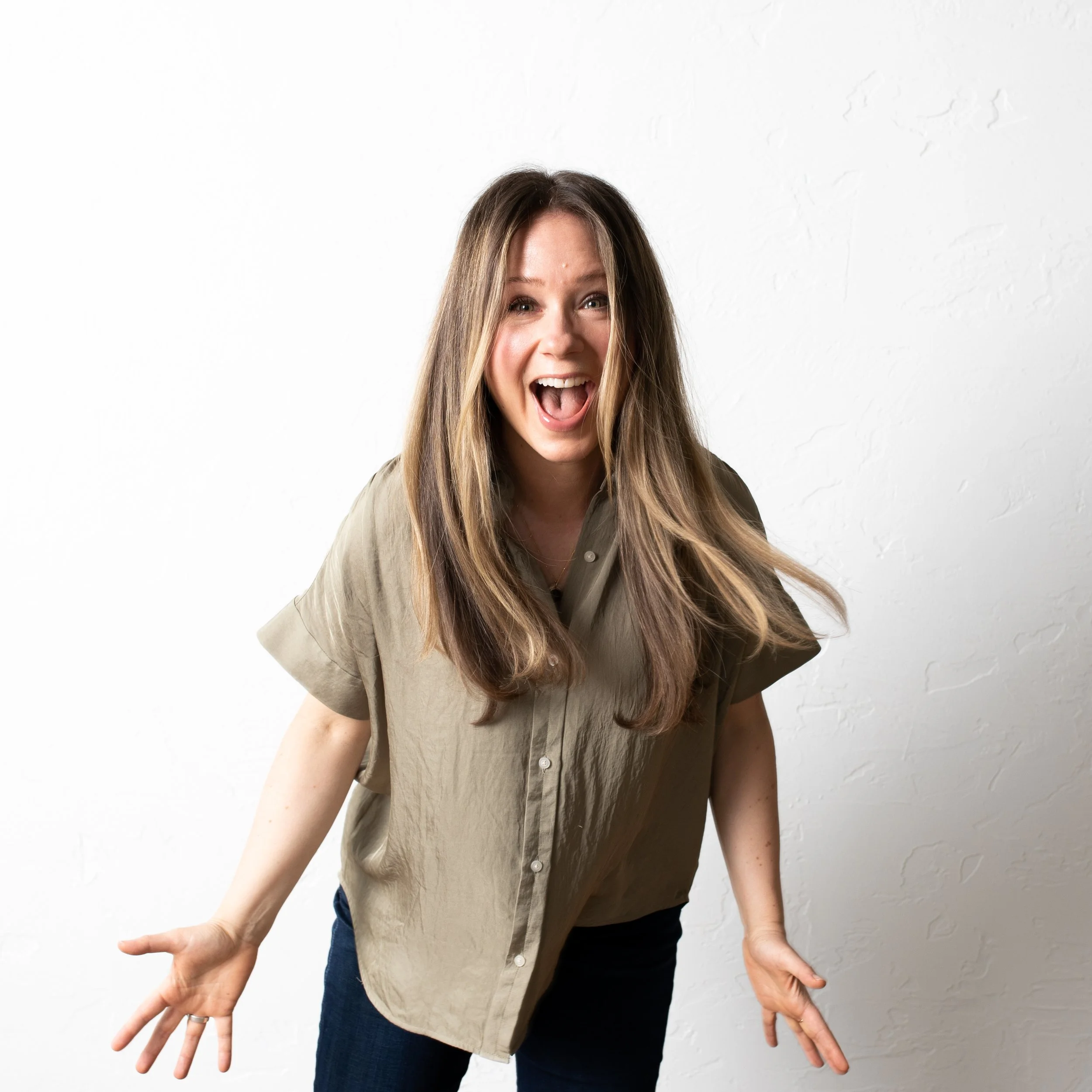 A woman with long brown hair smiling and gesturing with her hands against a white wall.