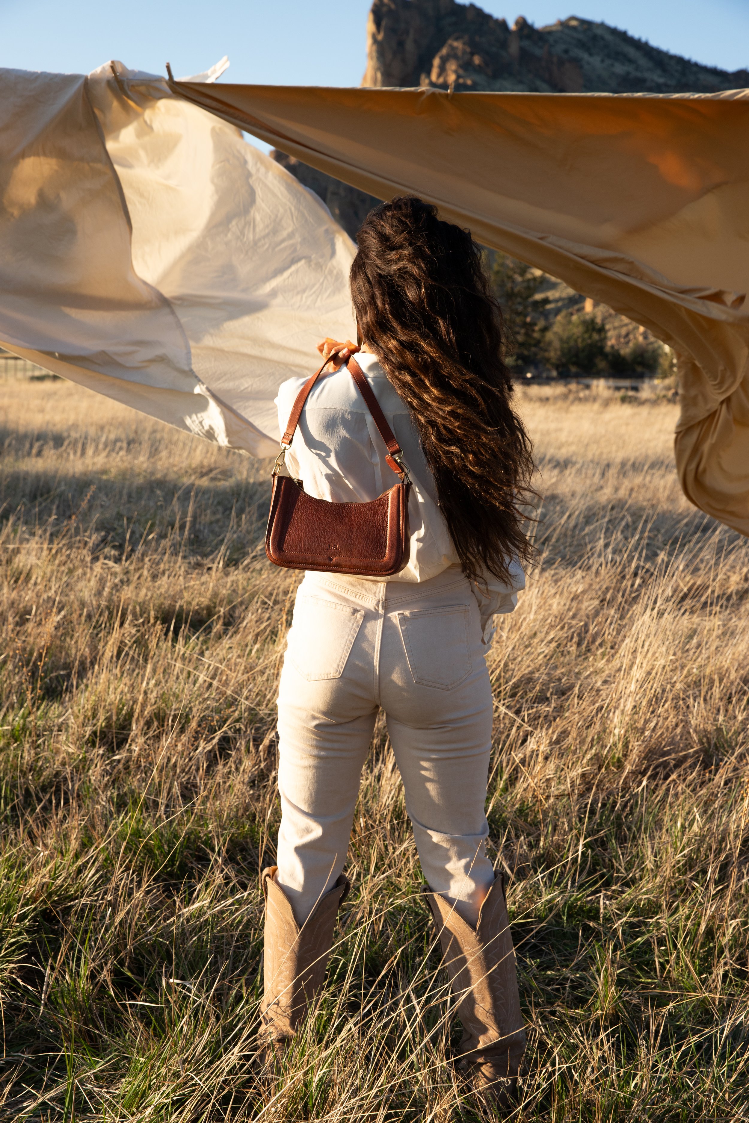 A woman with long, curly hair stands in a grassy field during sunset, holding a large fabric sheet or tent, with mountains in the background and a clear sky.