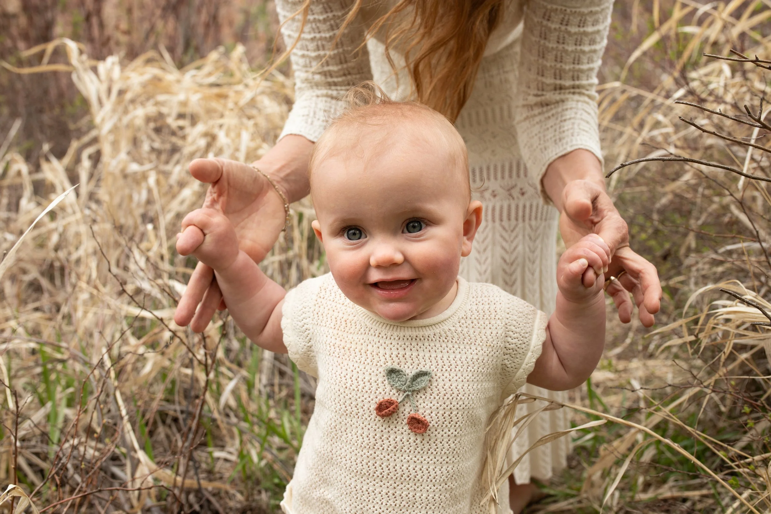 A smiling baby being supported by an adult's hands in a field of dried grass and plants.