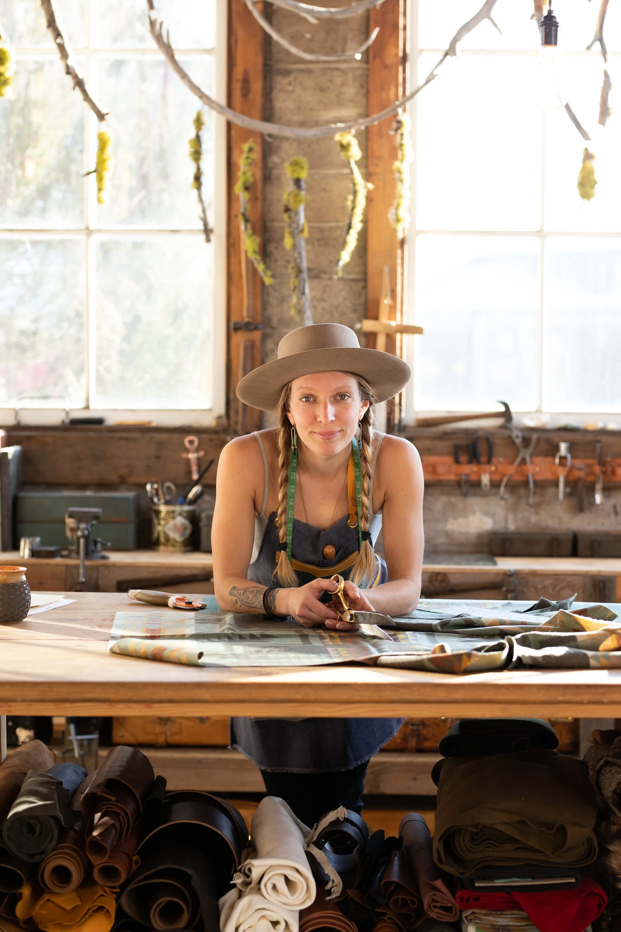 A woman in a wide-brimmed hat and apron working at a sewing table in a workshop with fabric rolls underneath and tools on the back wall, illuminated natural light from windows behind her.