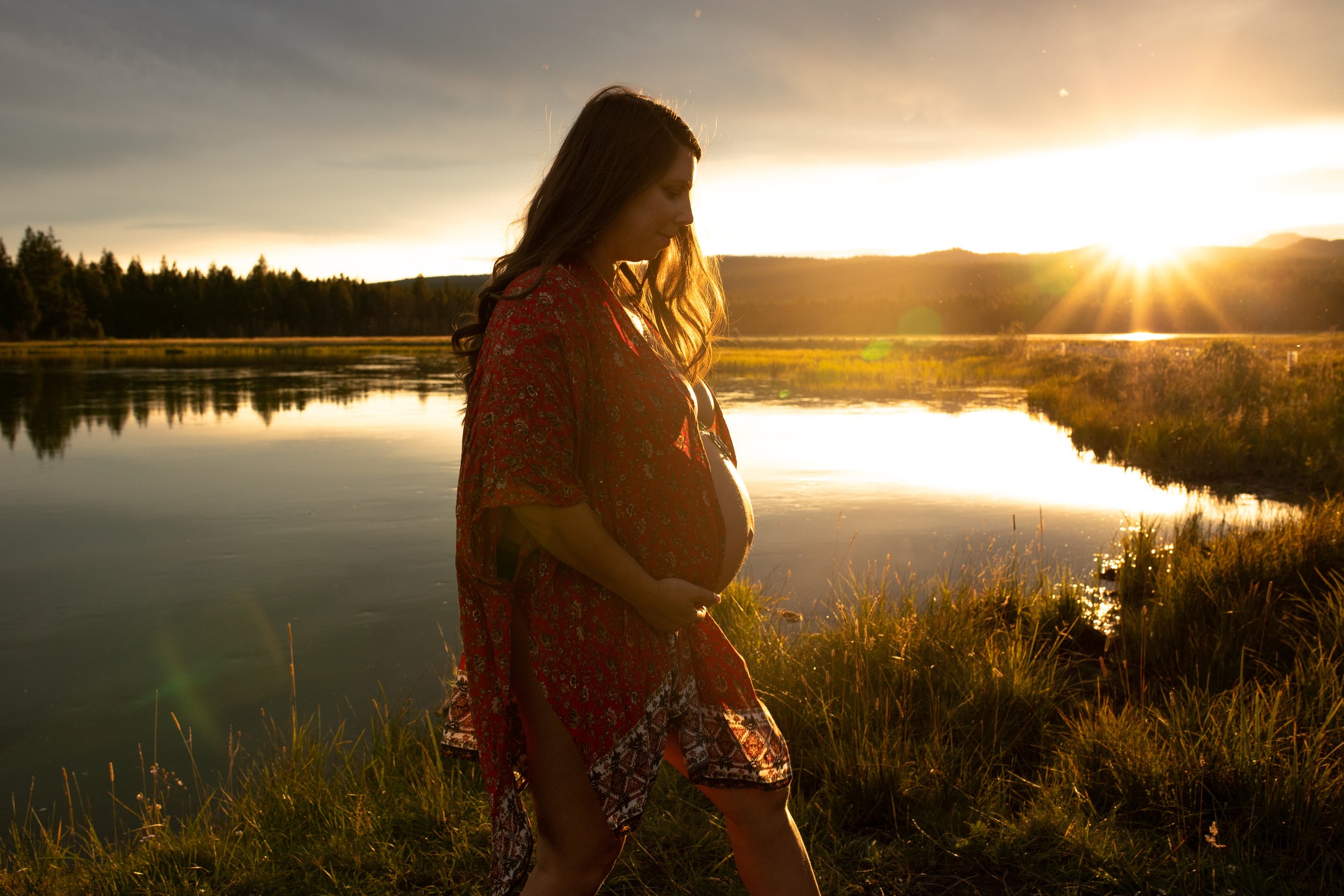 Pregnant woman standing by a lake at sunset in a red patterned robe, holding her belly.