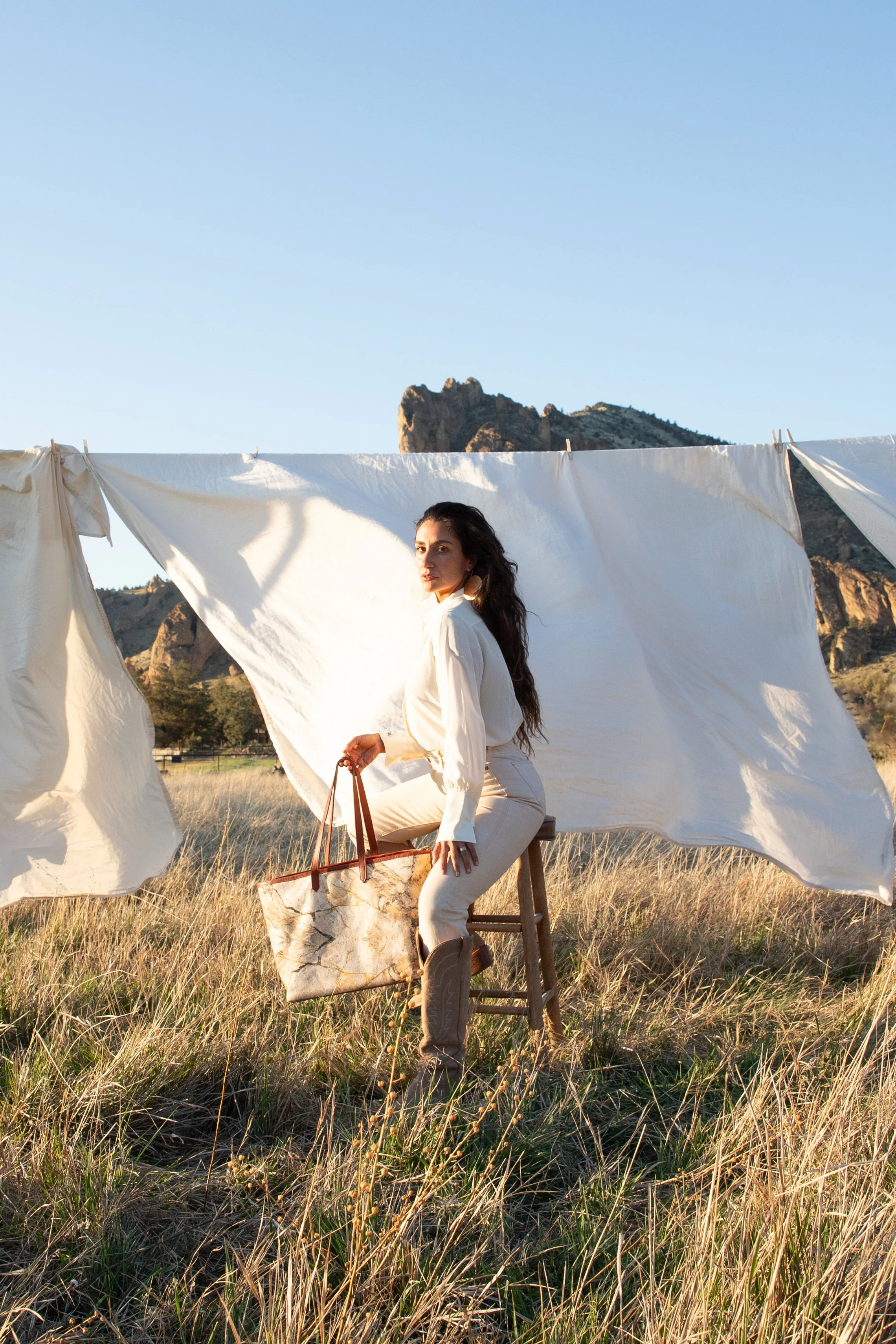 A woman with dark hair sitting on a stool in a grassy field, holding a tote bag, with white sheets hanging on a clothesline behind her, mountains in the background, and clear sky.