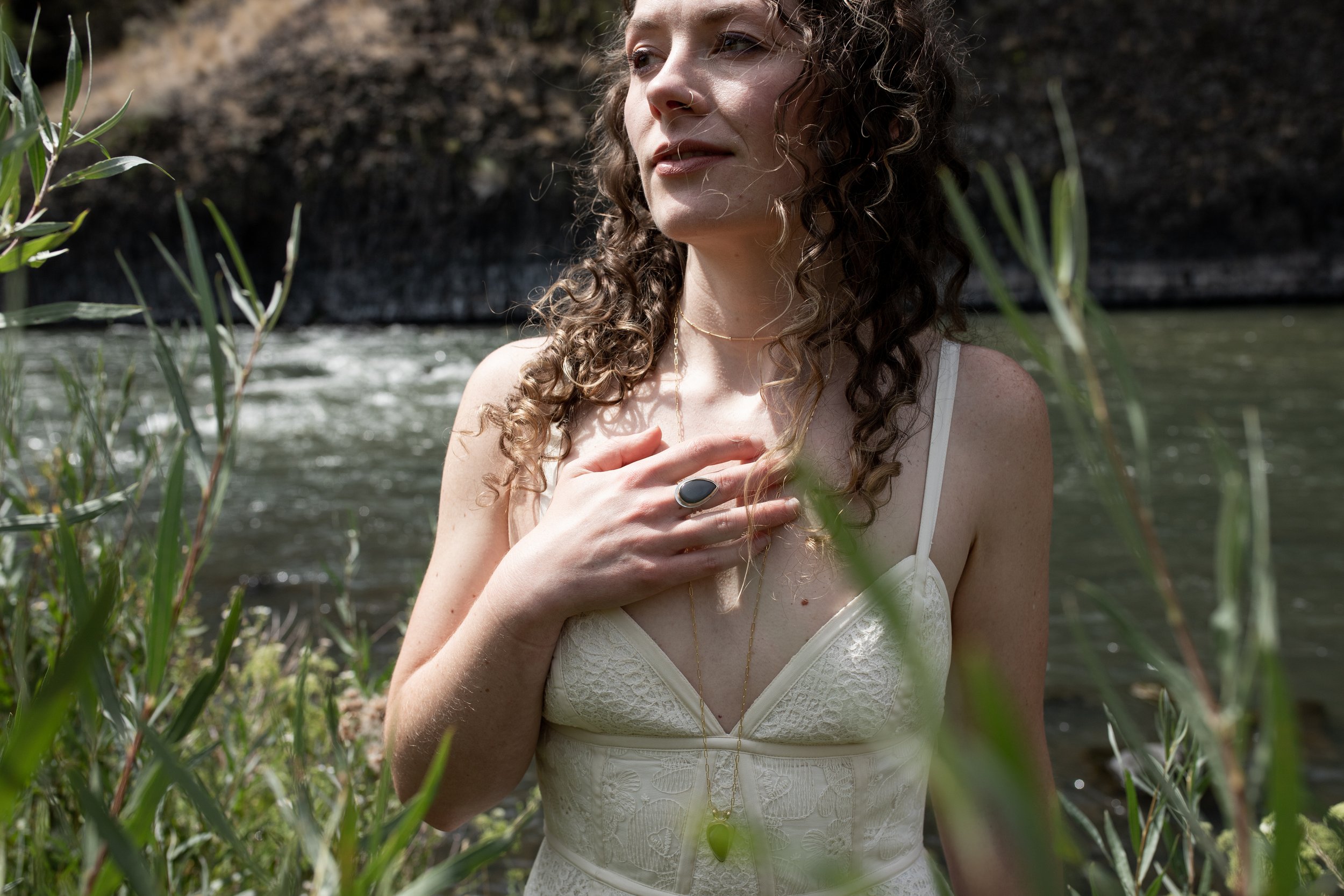 A woman with curly hair standing near a river, wearing a white dress and jewelry, with her hand on her chest.