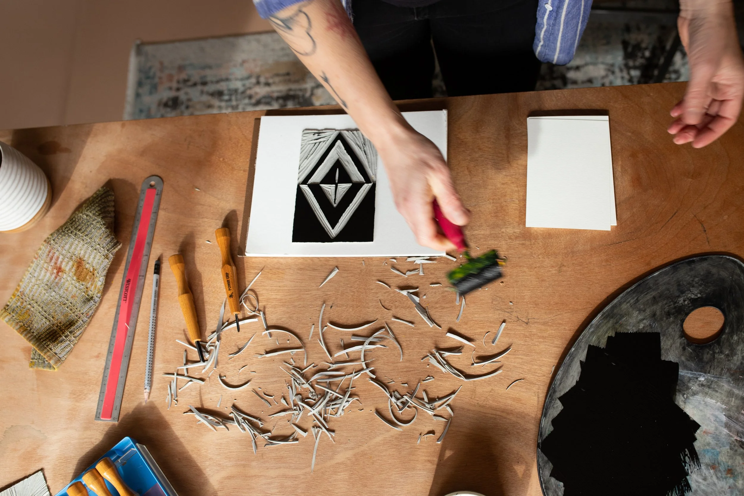 Person creating a stencil art of a diamond shape on paper, with tools and supplies scattered on a wooden table.