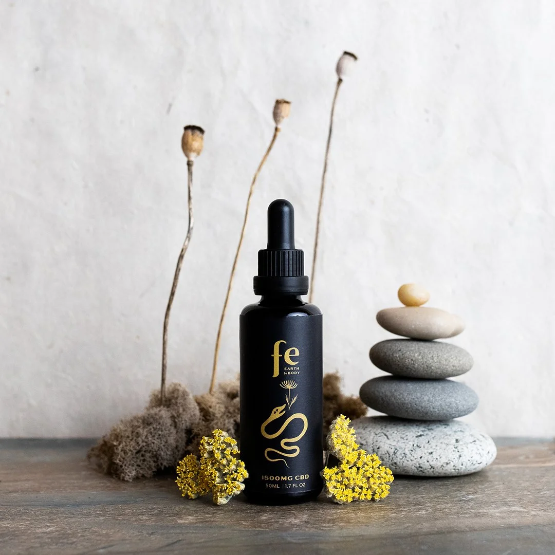 A black dropper bottle labeled FE Earth to Body with CBD and 1500mg on a wooden surface, surrounded by yellow flowers, stacked stones, dried plants, and moss with a plain beige wall background.
