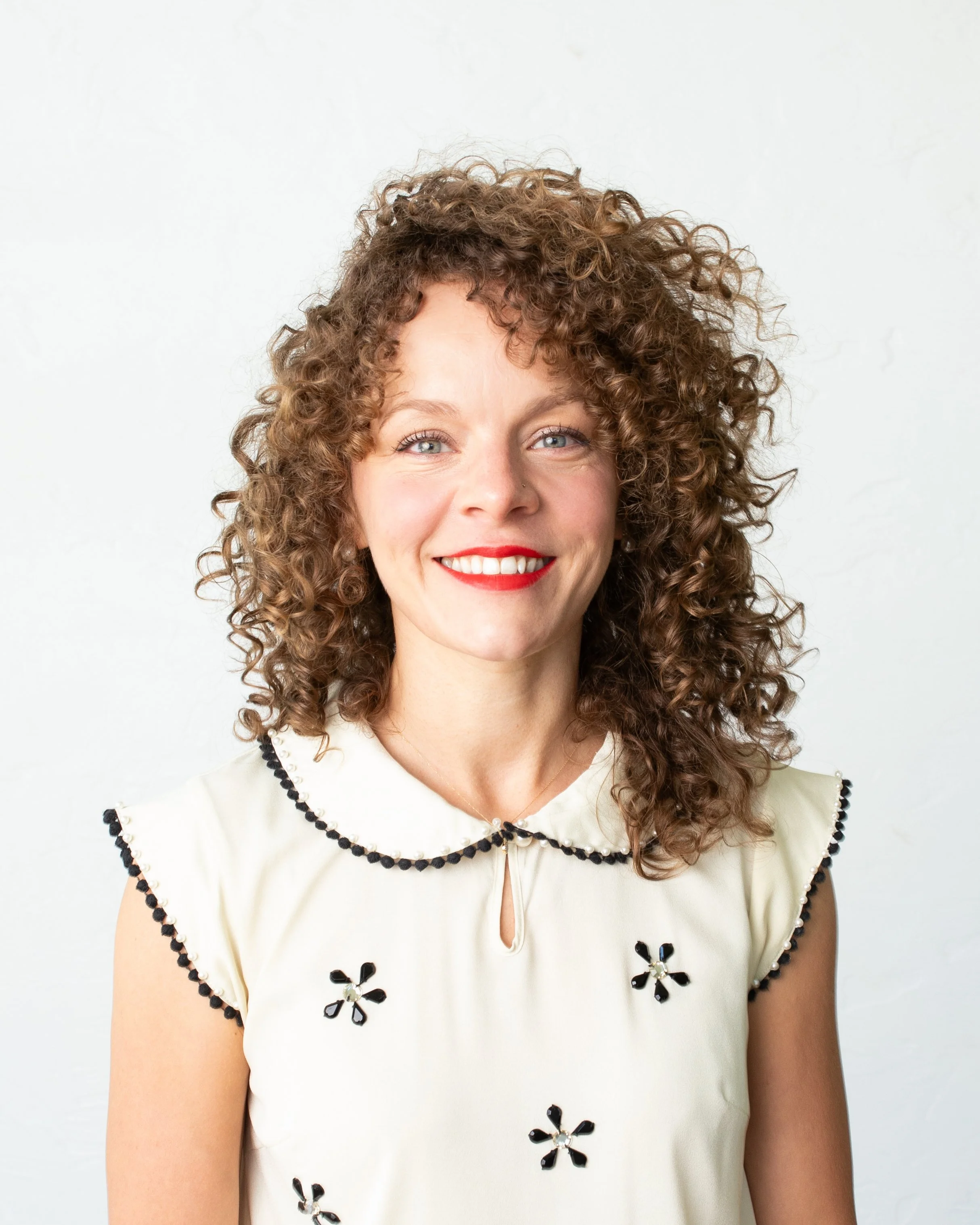 A woman with curly brown hair, blue eyes, and red lipstick smiles at the camera, wearing a cream sleeveless top with black flower embellishments and black bead accents on collar and sleeves against a white background.