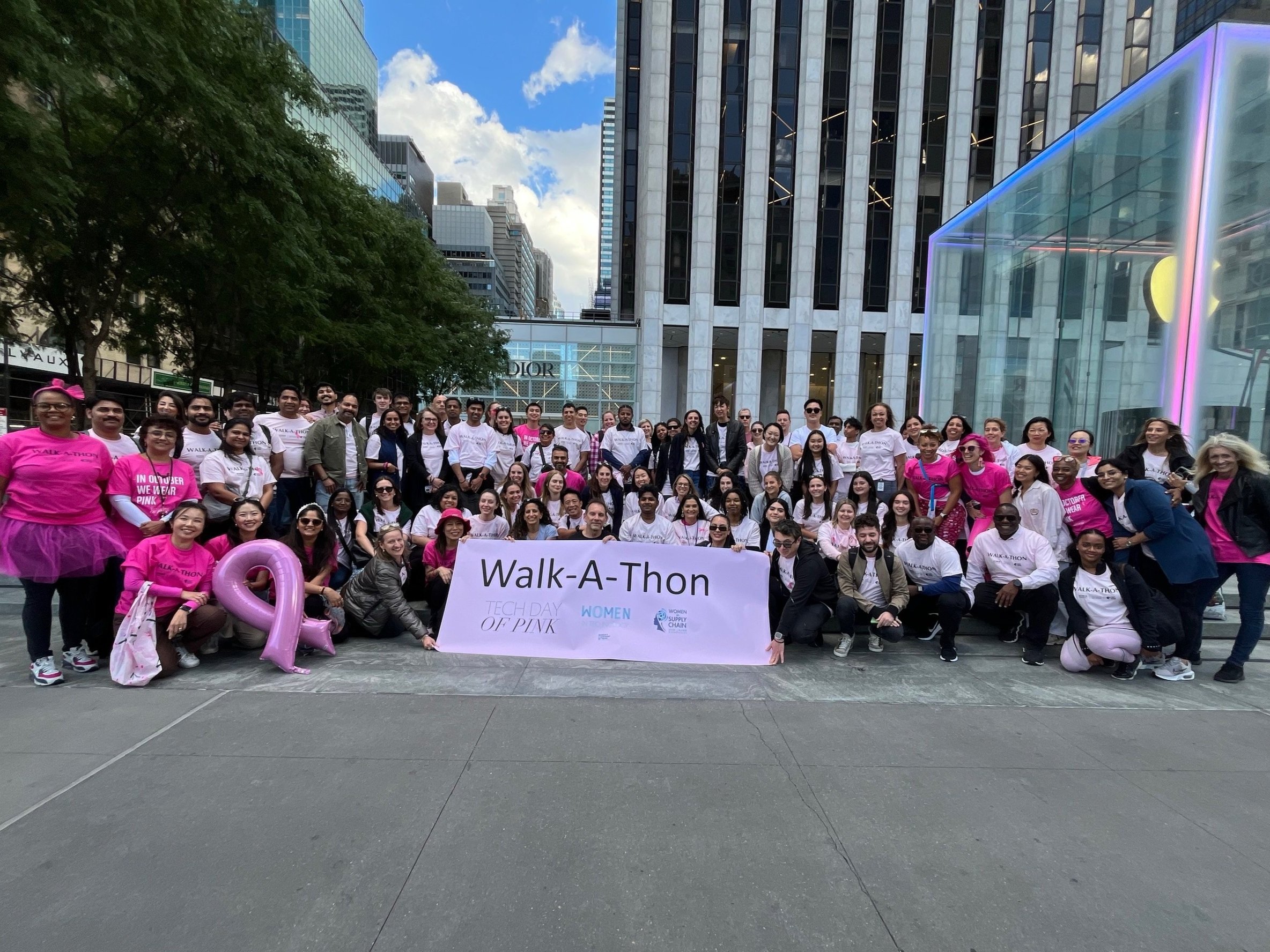 Group of people participating in a Walk-A-Thon event outdoors in an urban area, holding a banner that reads 'Walk-A-Thon Tech Day of Pink'. Some participants are wearing pink shirts and accessories, with city buildings in the background.