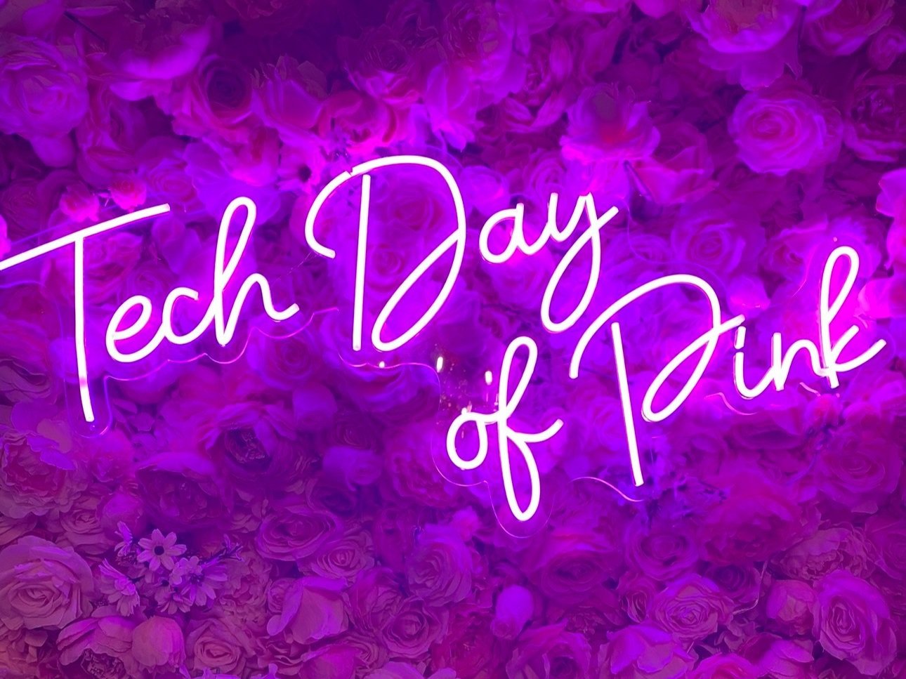 Neon sign that reads 'Tech Day of Pink' in bright pink outlined text, set against a background of purple-pink flowers.