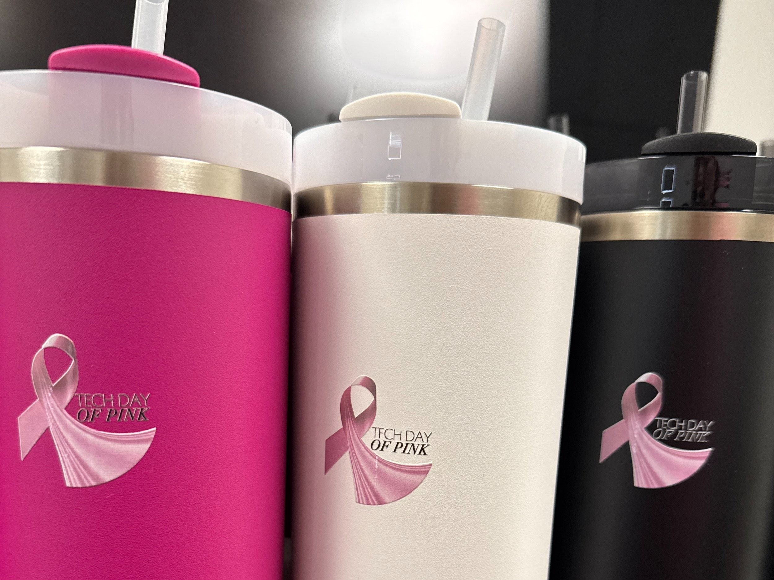 Three insulated tumblers with pink, white, and black colors, each with a breast cancer awareness ribbon and 'Tech Day of Pink' text, and straws on top.