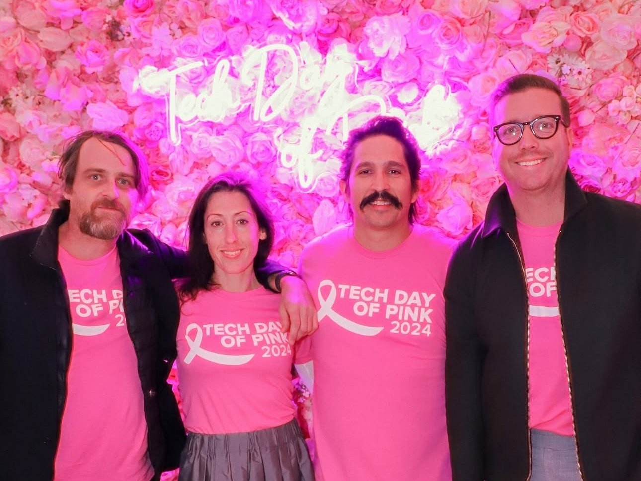 Four people standing together at a pink-themed event, wearing pink T-shirts with the text "Tech Day of Pink" and a pink ribbon logo, against a pink floral background with neon lighting.
