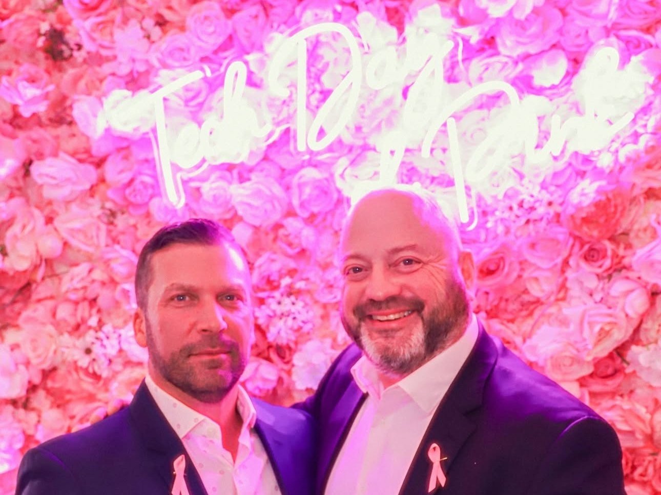 Two men in suits standing in front of a pink flower wall with a neon sign that reads "Tech Day of Pink" and pink ribbon pins on their lapels.