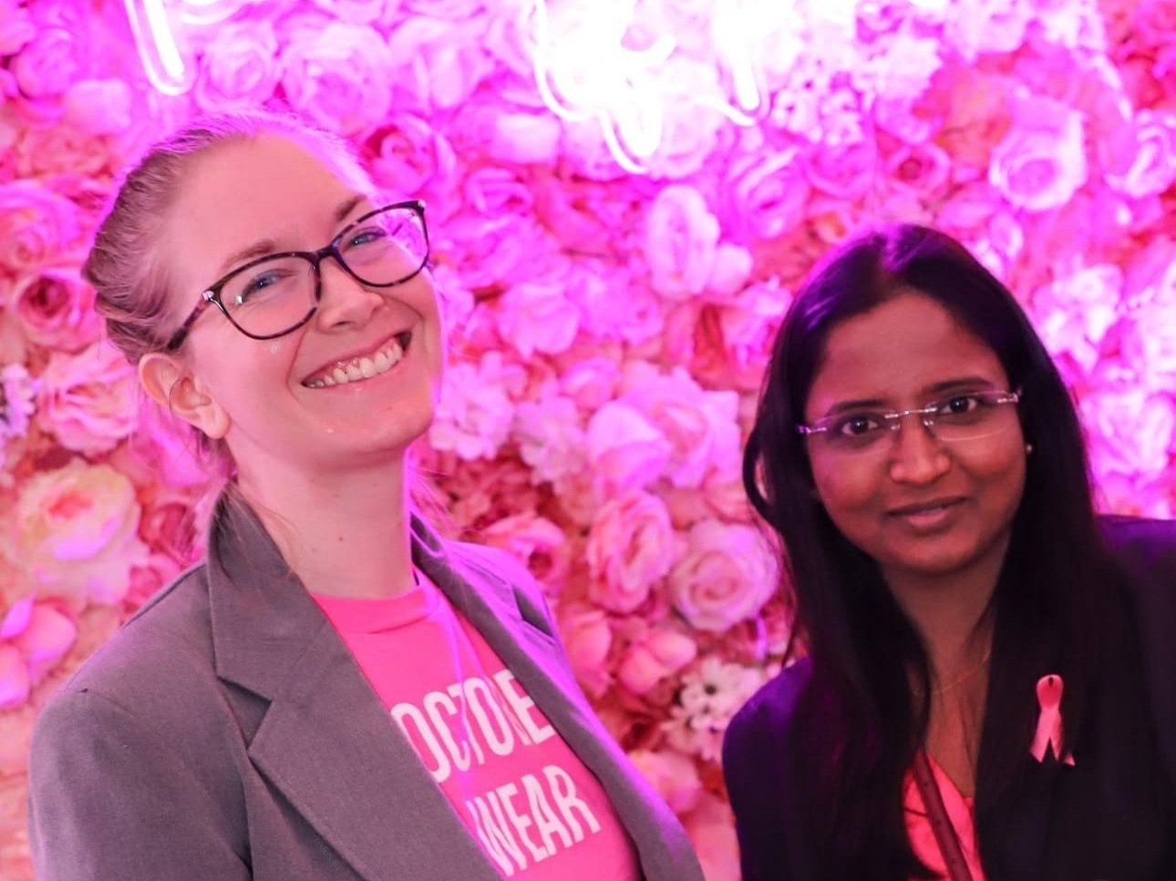 Two women standing in front of a pink flower wall; one woman has blonde hair, glasses, and is smiling, wearing a gray blazer and pink shirt; the other woman has dark hair, glasses, and a pink ribbon pin, smiling gently, wearing a dark blazer.