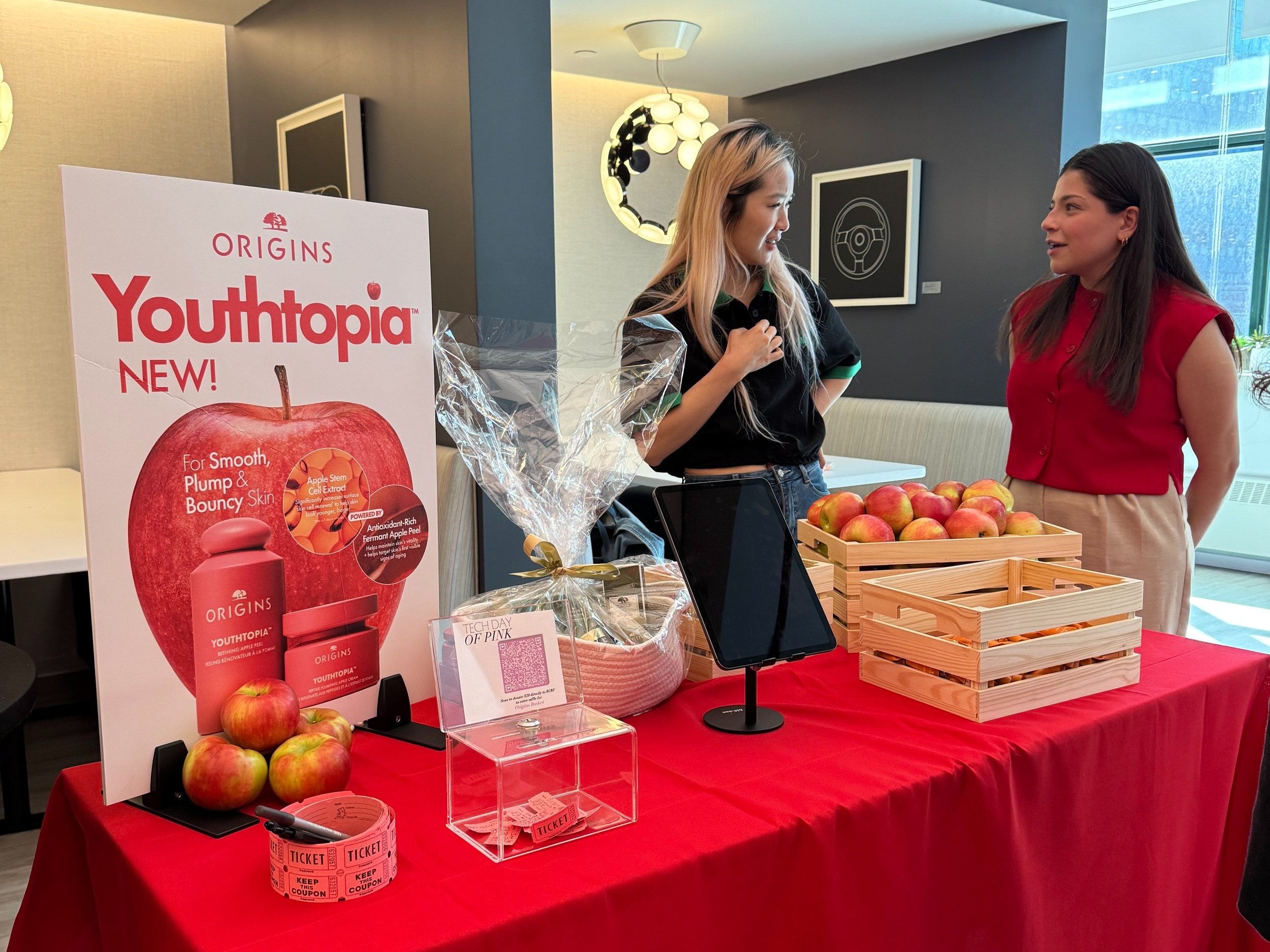 Two women at a table selling apples and promoting Youthtopia skincare products, with a large Youthtopia poster, wooden crates of apples, tickets, and a tablet.
