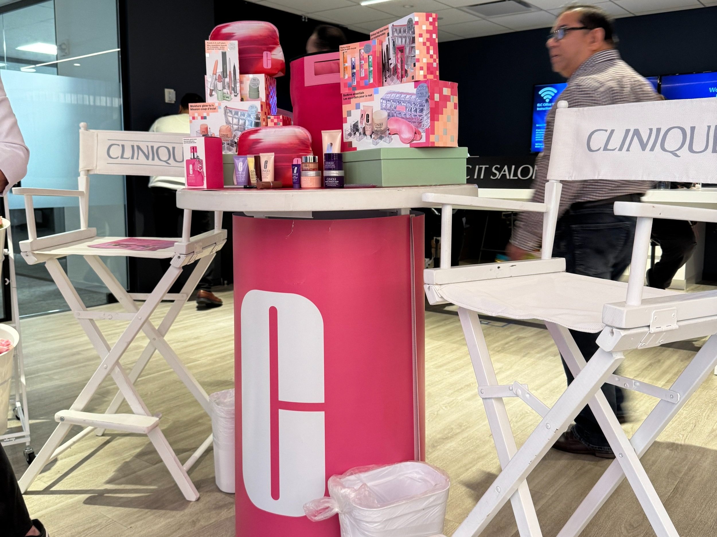 Clinique skincare display at a promotional event with two white folding chairs, pink and green packaging, and product samples on a small round table.